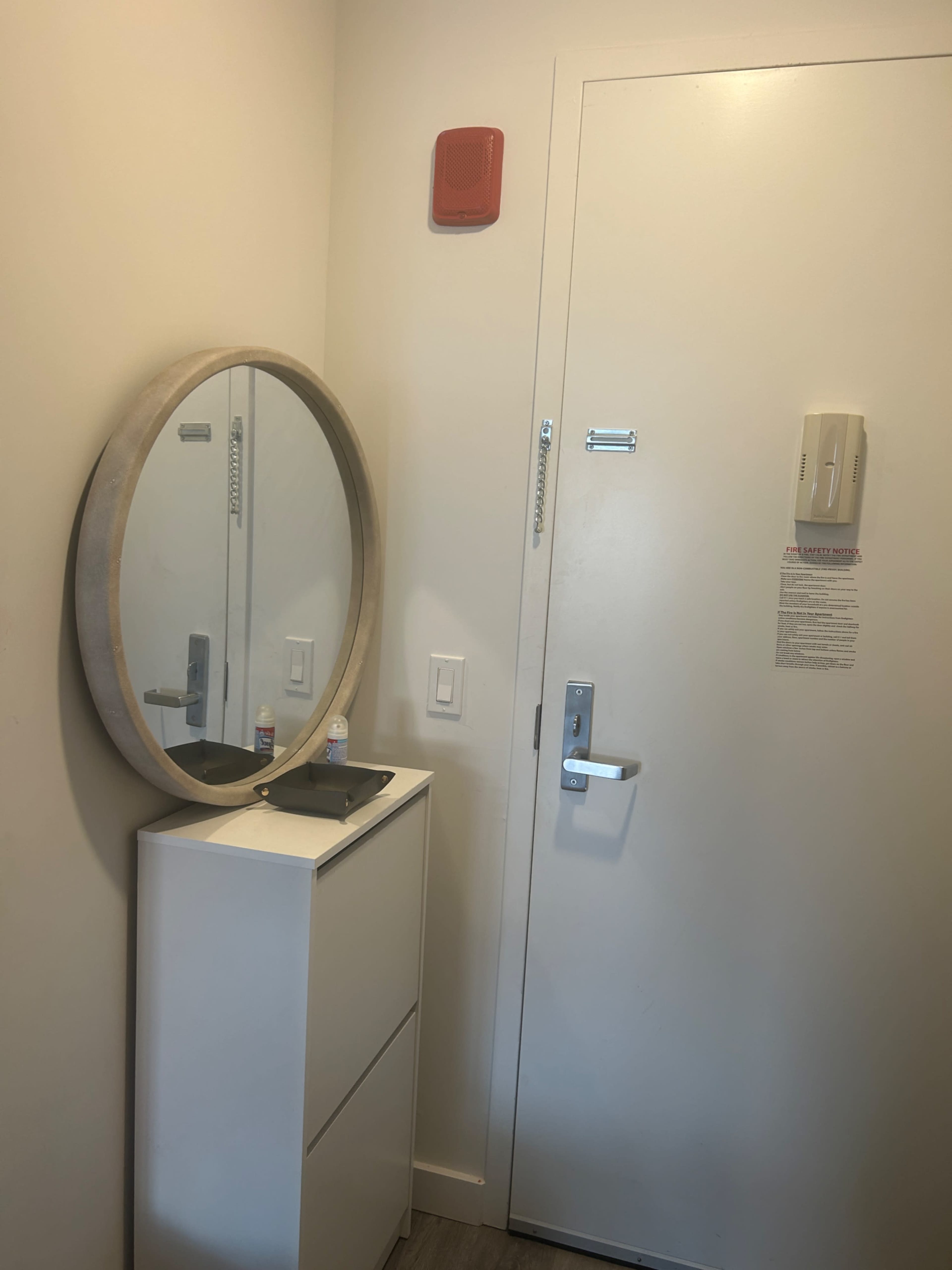 The image shows a small entryway featuring a round mirror above a white dresser and a door with an electronic lock and a fire alarm.