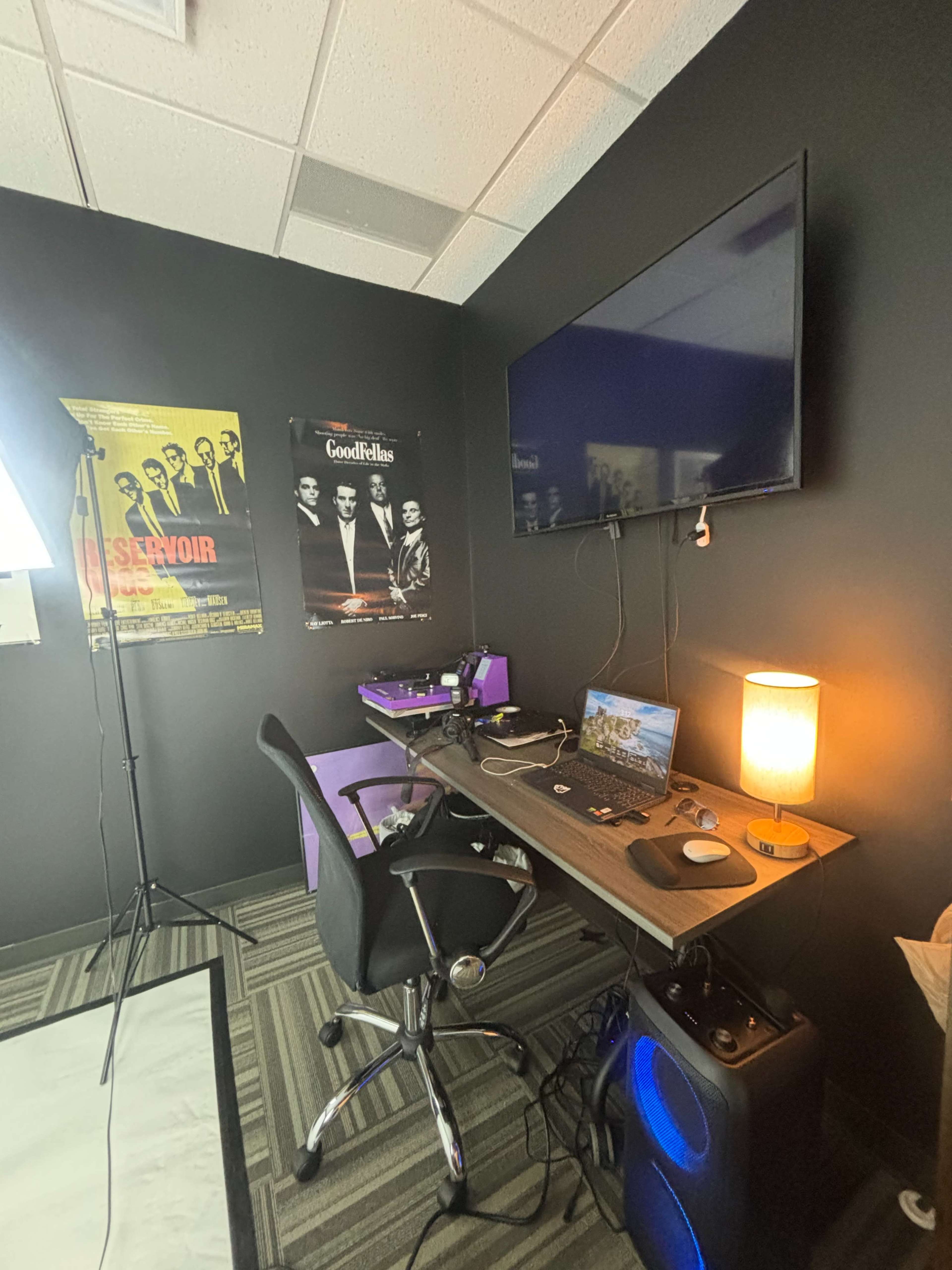 The image shows a workspace with a desk, computer setup, lamp, and posters on the wall in a dark-colored room.