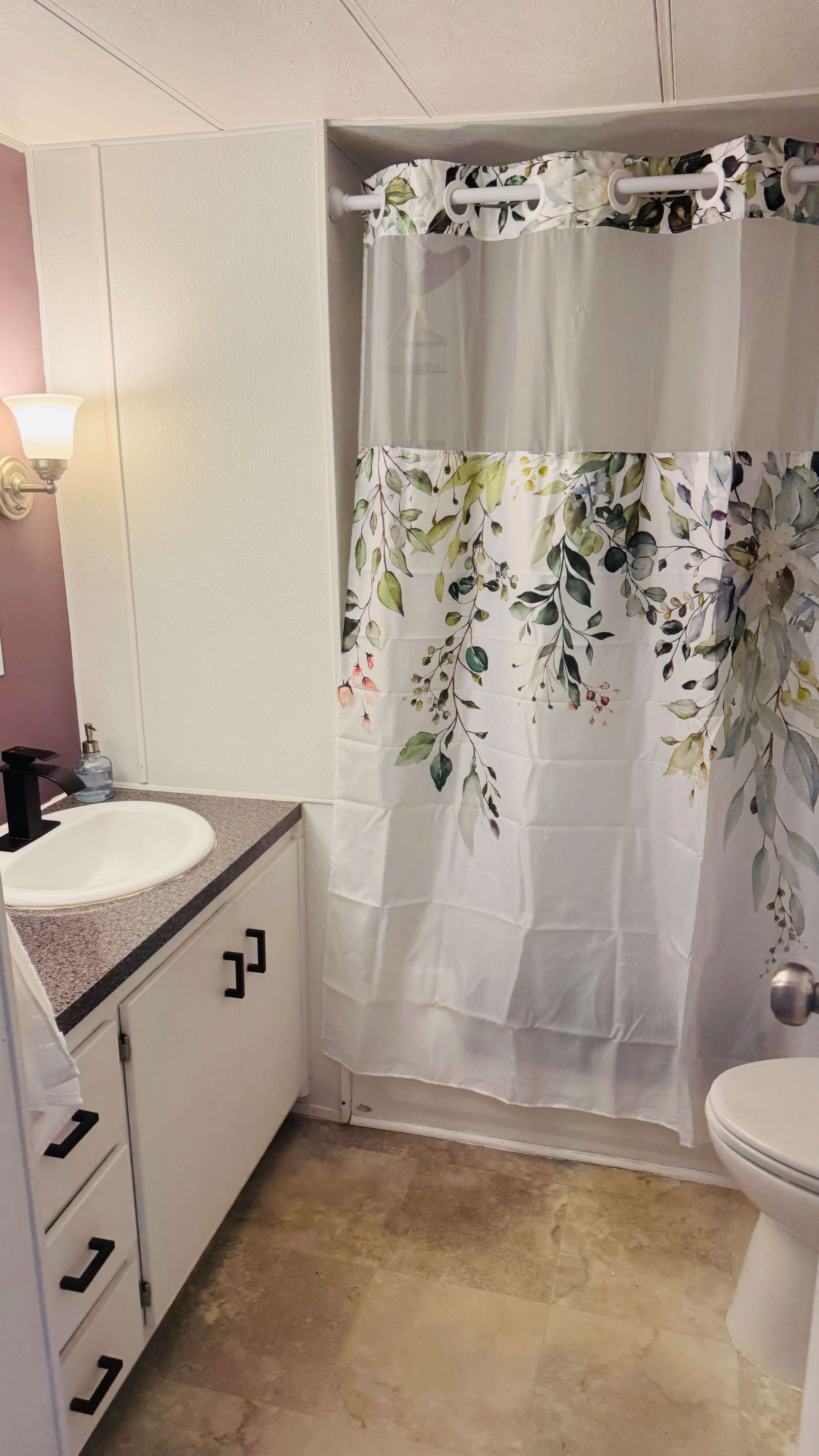 The image shows a bathroom with a sink, a shower enclosed by a floral curtain, and a toilet in a modest setting.