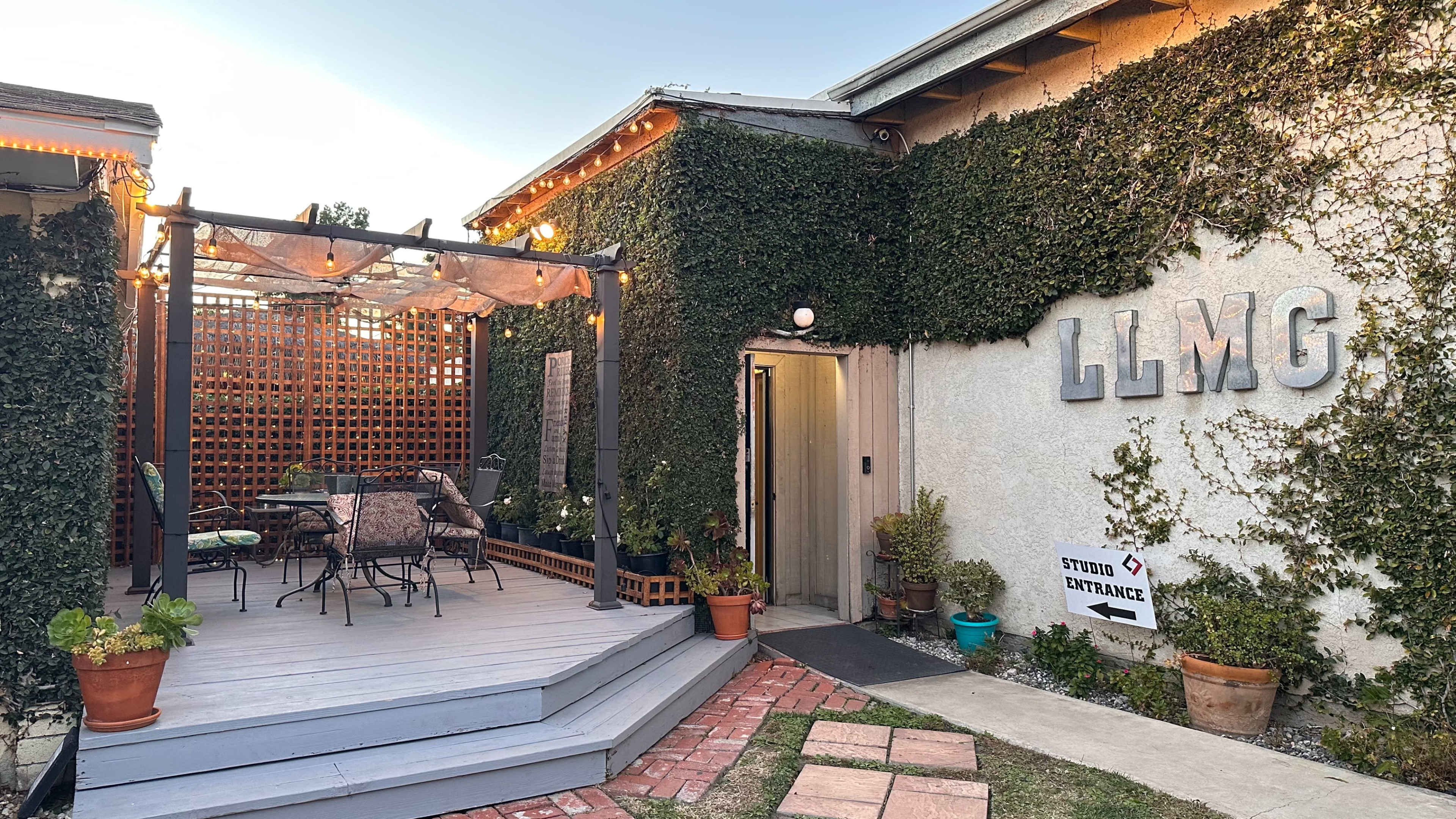 A building with an outdoor seating area, featuring a pergola covered in vines, and a sign indicating the studio entrance.