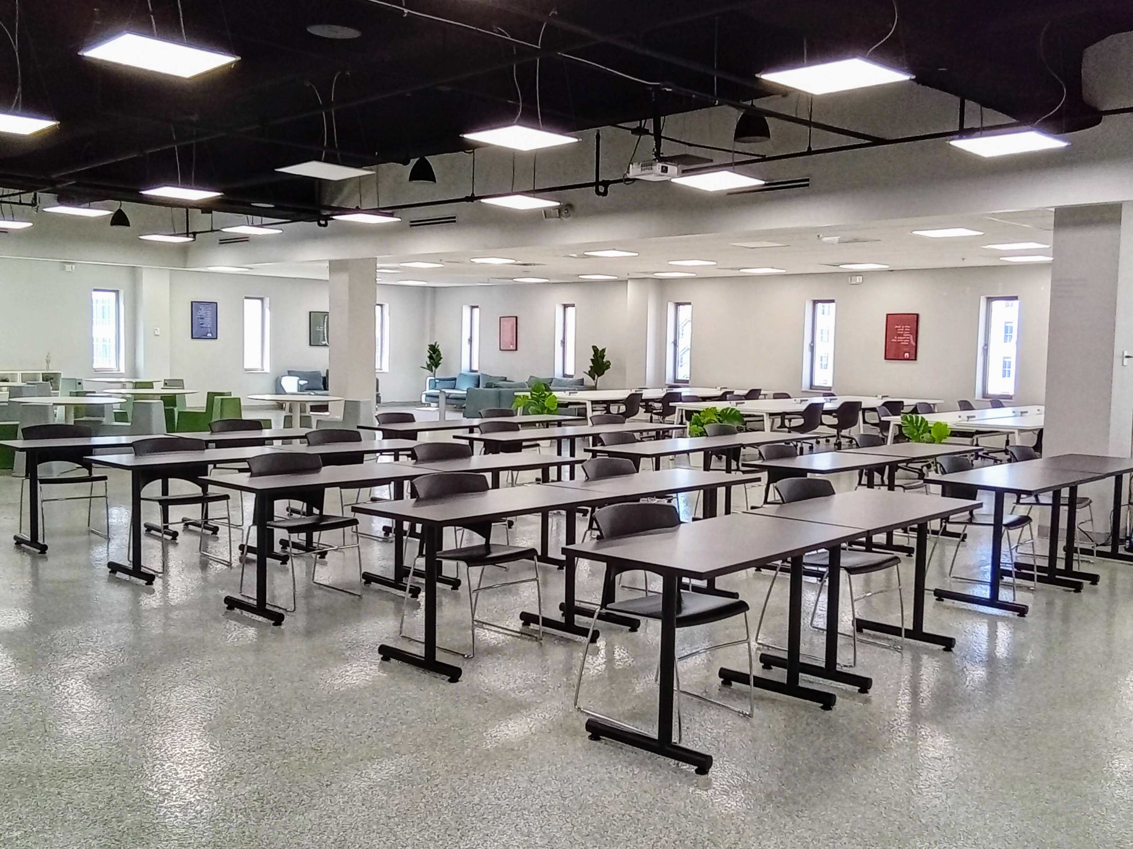 The image shows a spacious classroom with multiple rows of tables and chairs, set against a backdrop of large windows and green plants.