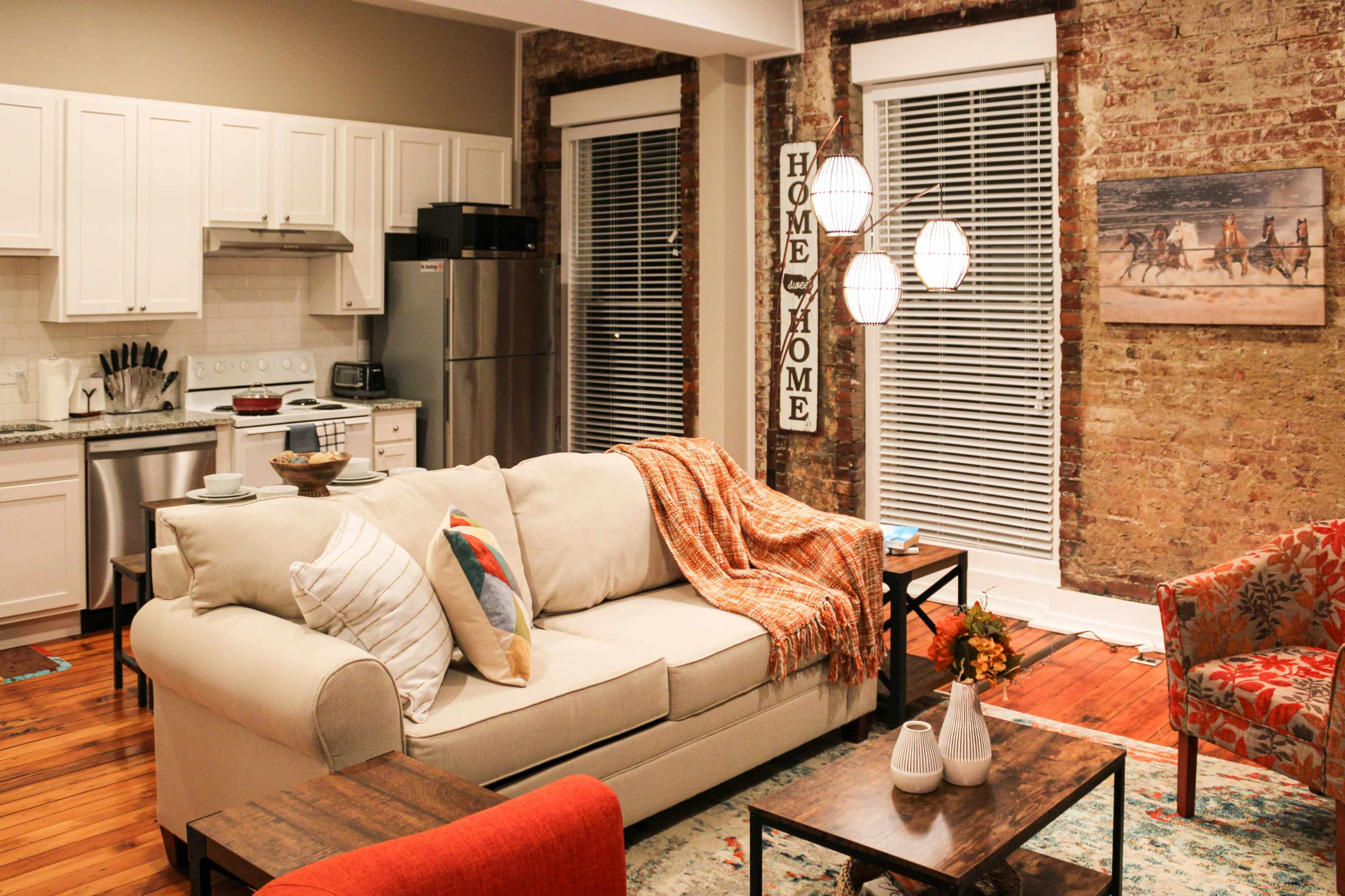 A modern living room with a light-colored sofa, a wooden coffee table, and a kitchen area in the background, featuring exposed brick walls and large windows with blinds.