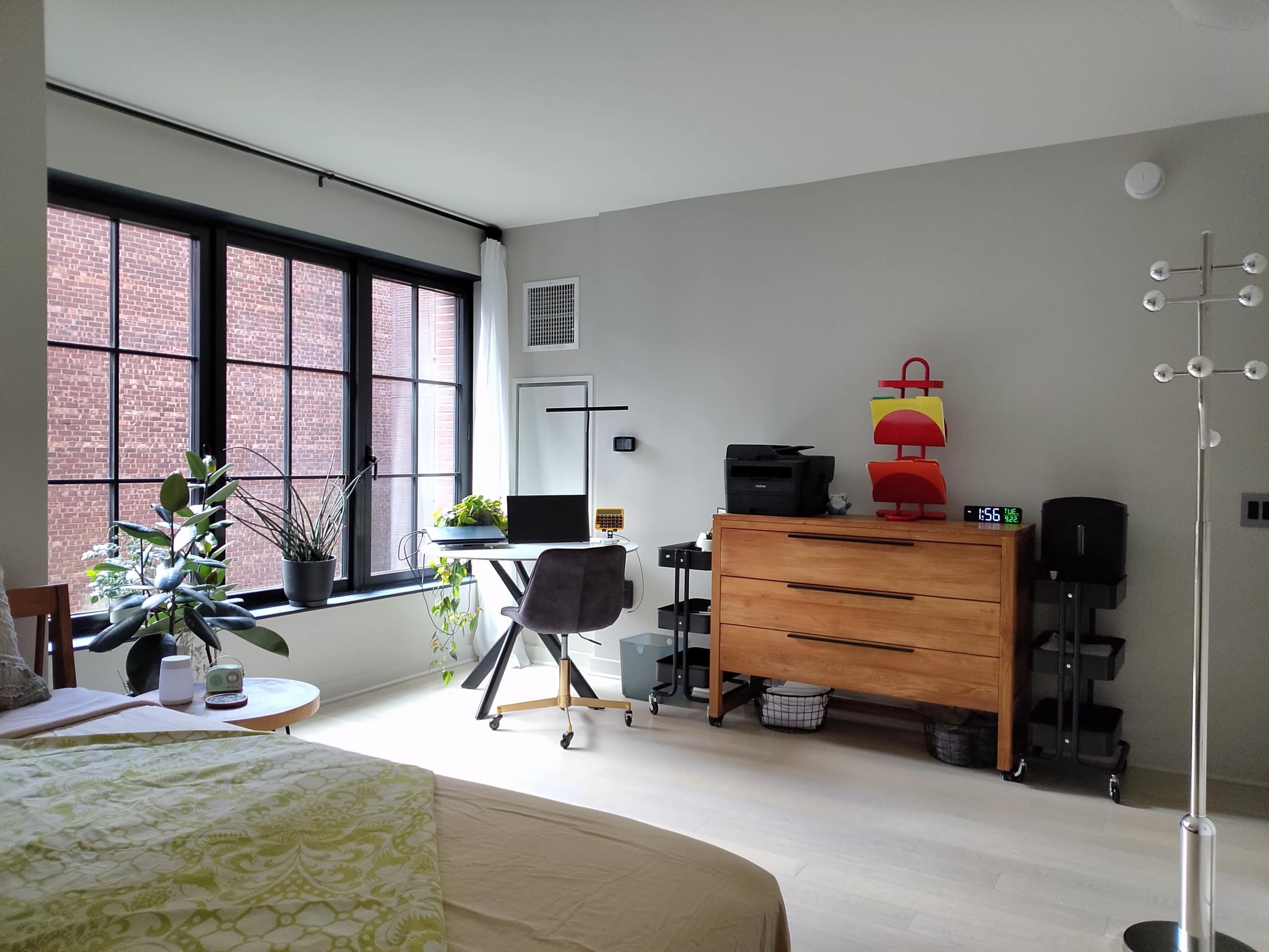 The image shows a light-colored room with a bed in the foreground, a wooden dresser, a workspace with a desk and plants, and large windows allowing natural light to enter.