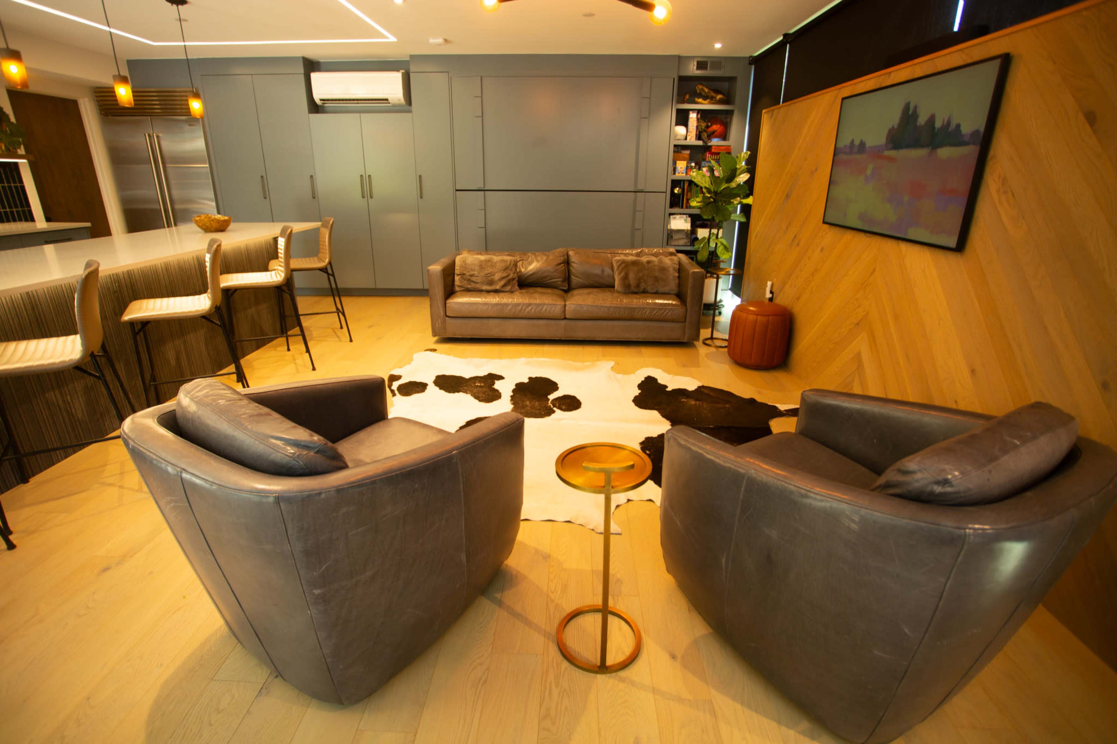 The image shows a modern living room featuring a sofa and two armchairs arranged around a cowhide rug, with a kitchen area and decorative shelves in the background.
