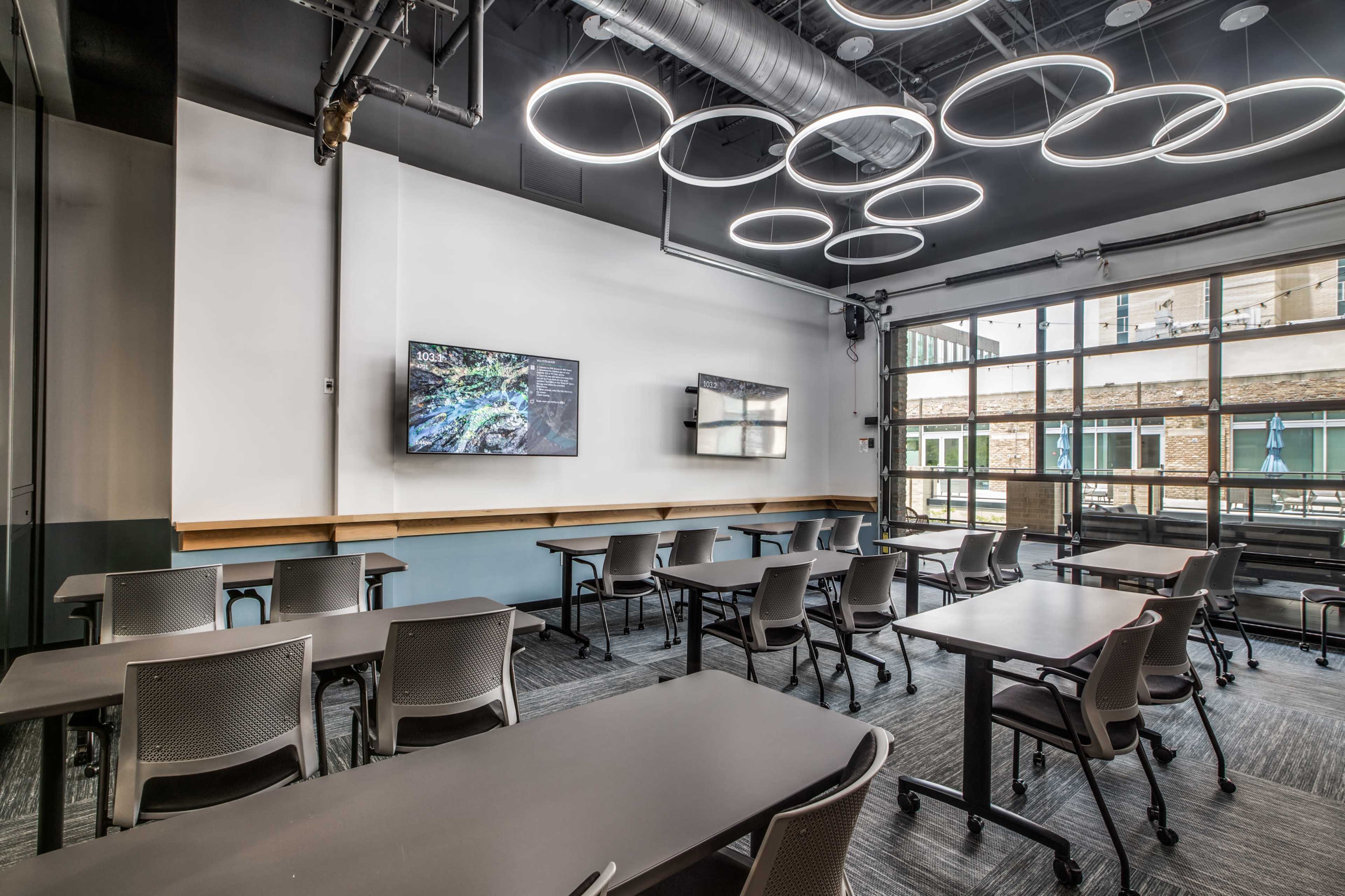 The image shows a modern classroom with several movable desks arranged for collaboration, featuring two wall-mounted screens and circular light fixtures overhead.