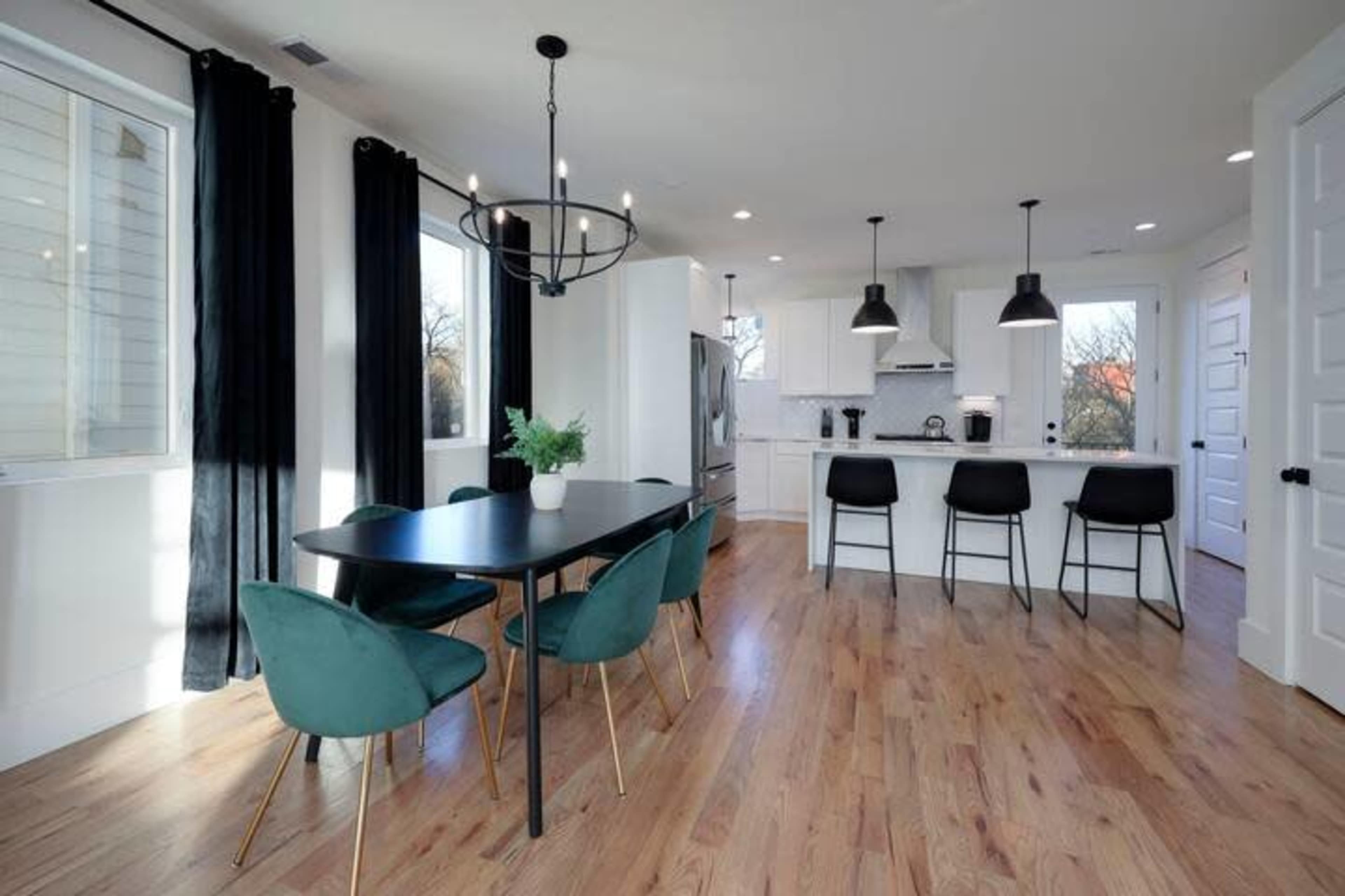 The image shows a modern kitchen and dining area featuring a rectangular black table with green chairs, a kitchen island with bar stools, and large windows allowing natural light.