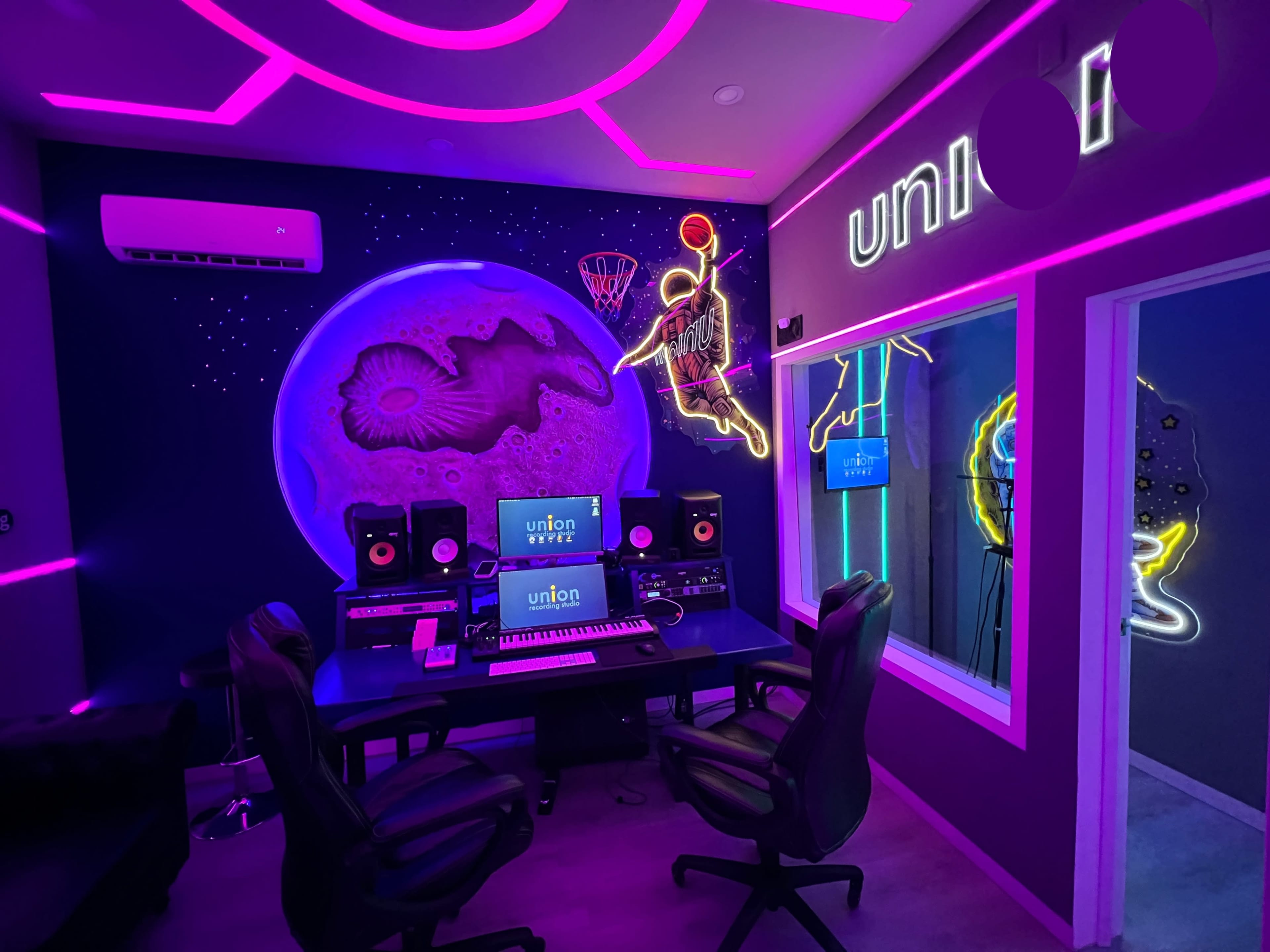 The image shows a modern music studio featuring a neon-lit interior with a large mural of a basketball player and a moon backdrop, accompanied by a central mixing desk and sound monitors.
