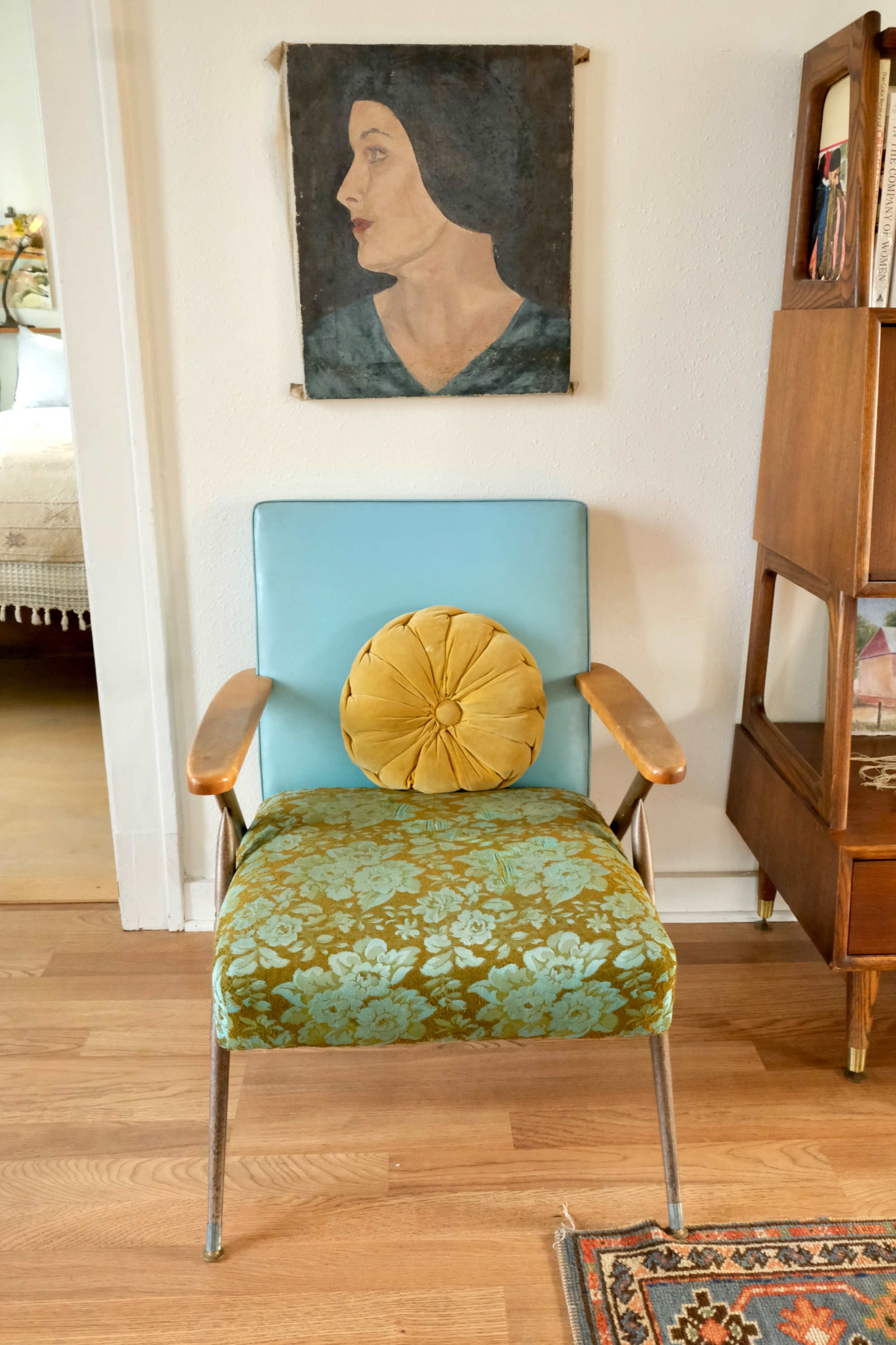 A vintage chair with patterned upholstery and a round cushion sits in front of a portrait of a woman mounted on the wall.