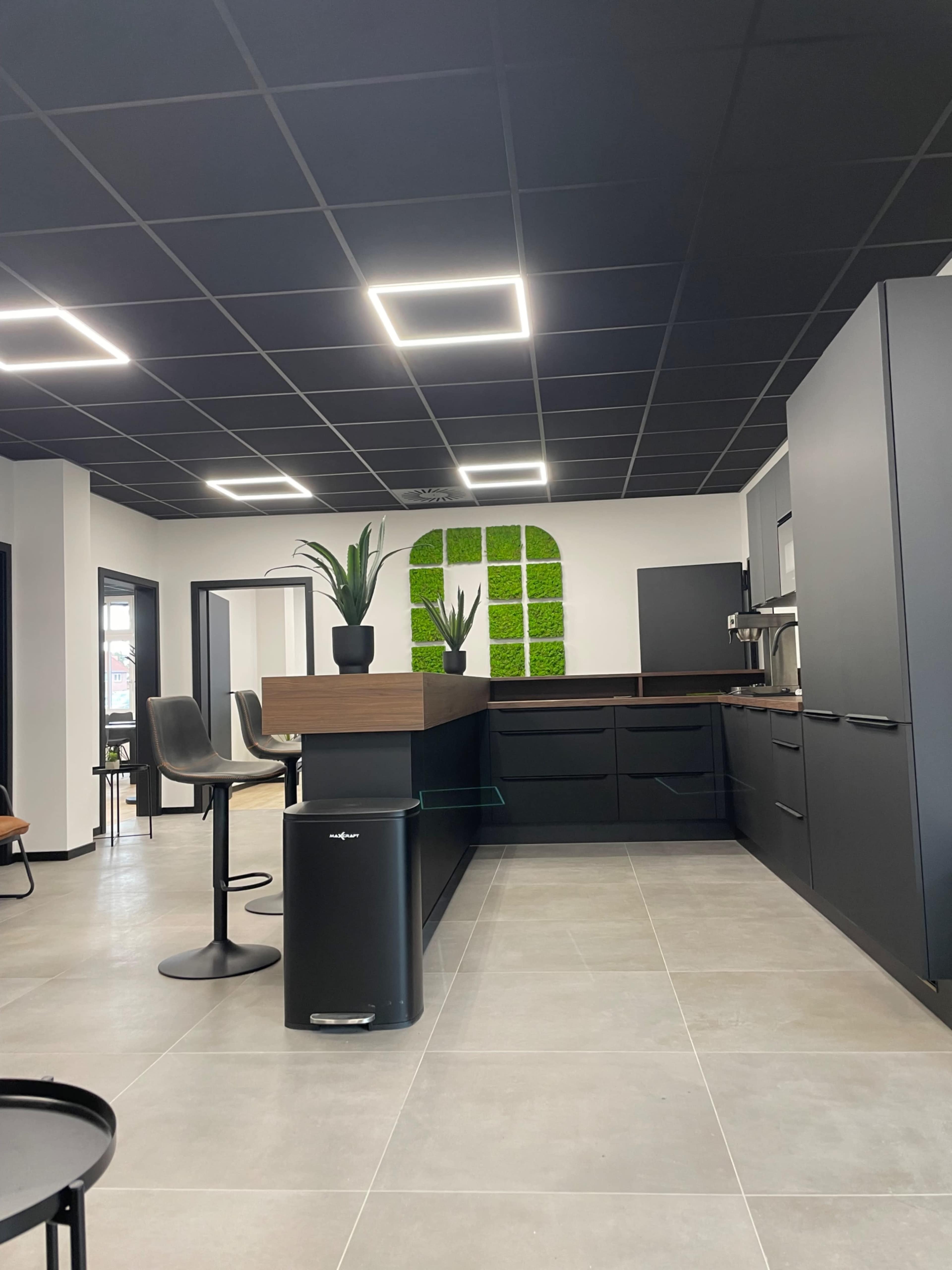The image depicts a modern kitchen with a black ceiling, sleek cabinetry, a central island with bar stools, and a decorative green moss wall.