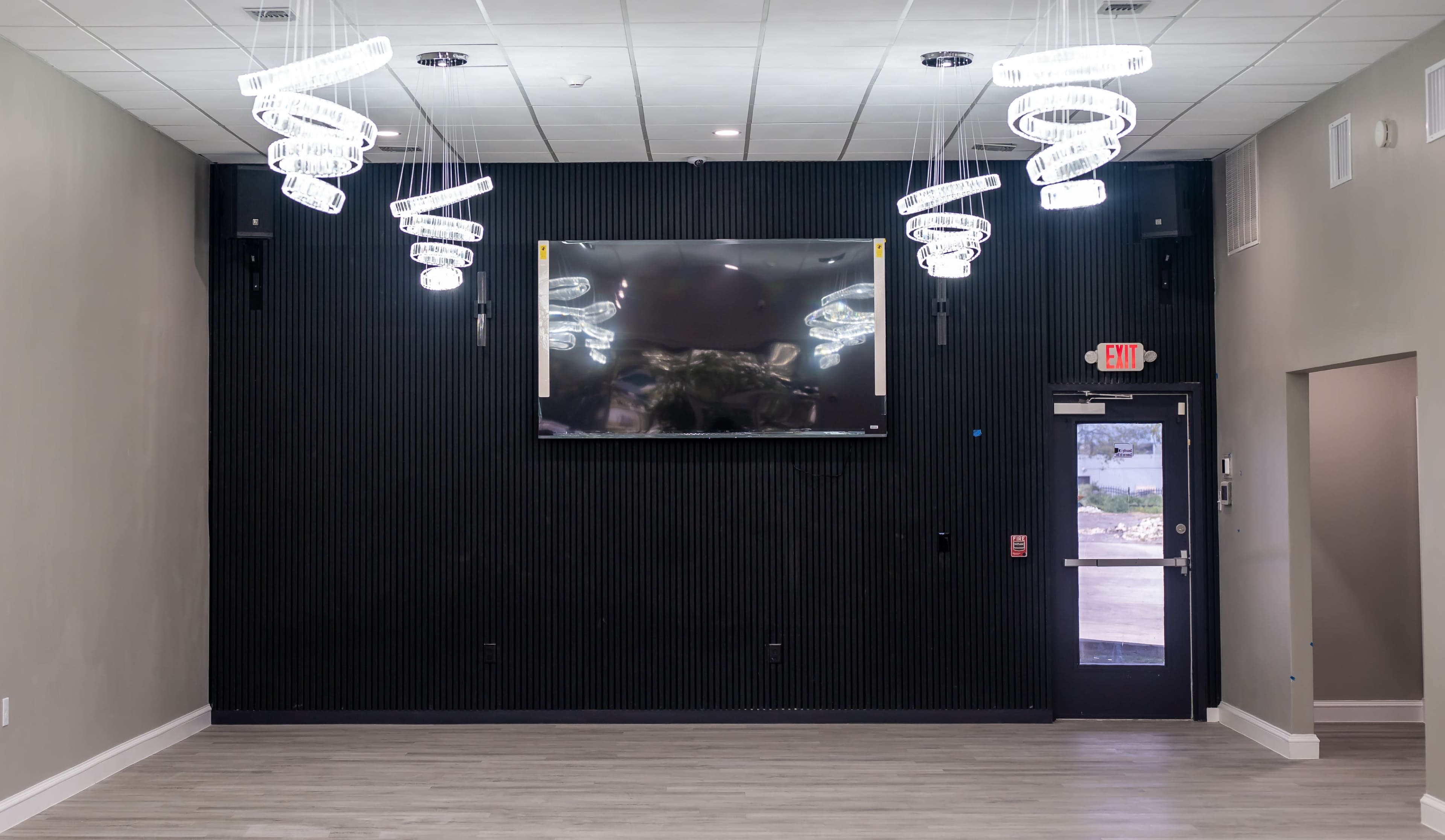 The image shows a modern interior space featuring a large flat-screen TV mounted on a black accent wall with decorative light fixtures hanging from the ceiling.