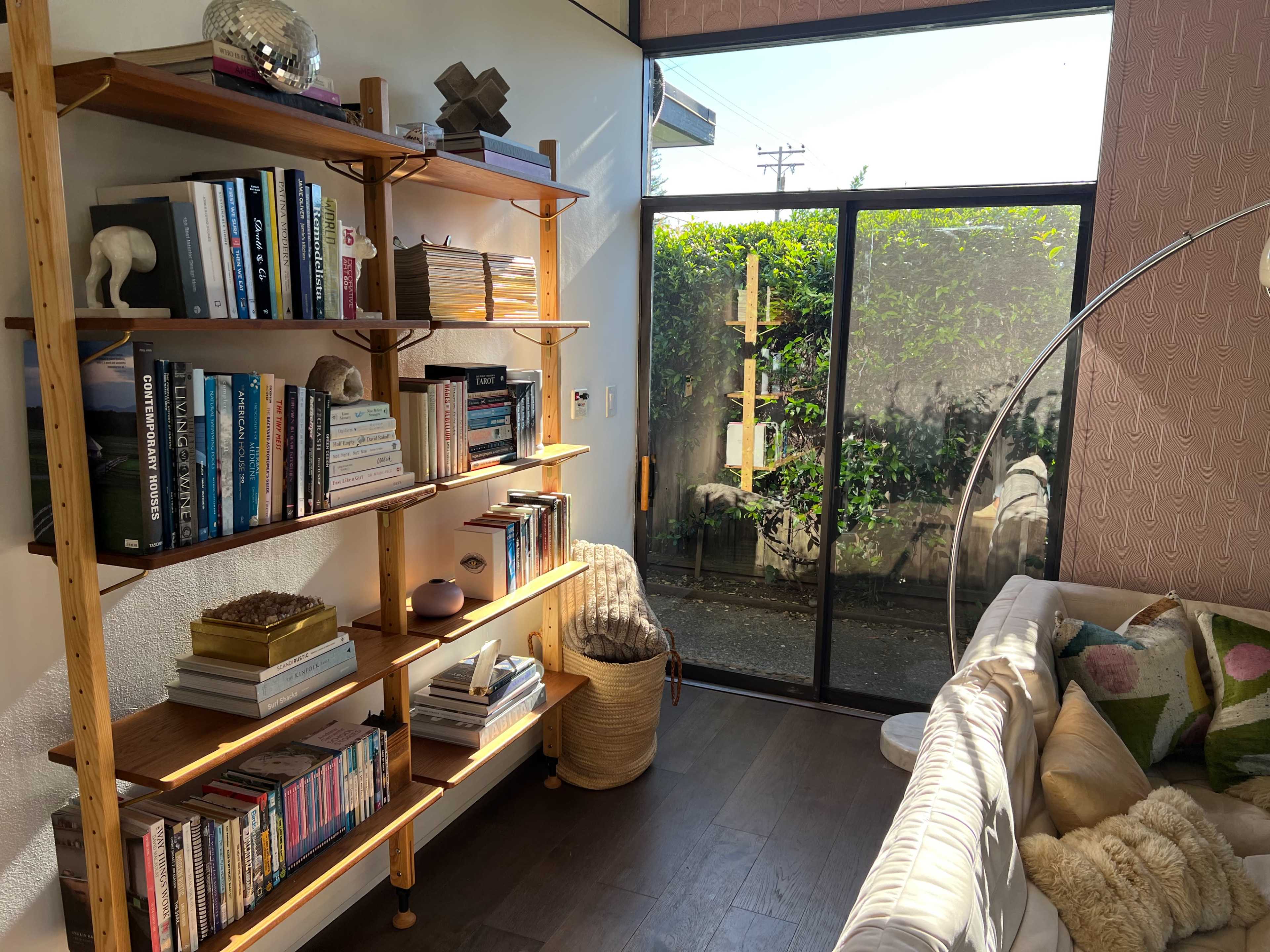The image shows a bright room with a wooden bookshelf filled with books, a cozy couch, and large windows overlooking a green outdoor space.