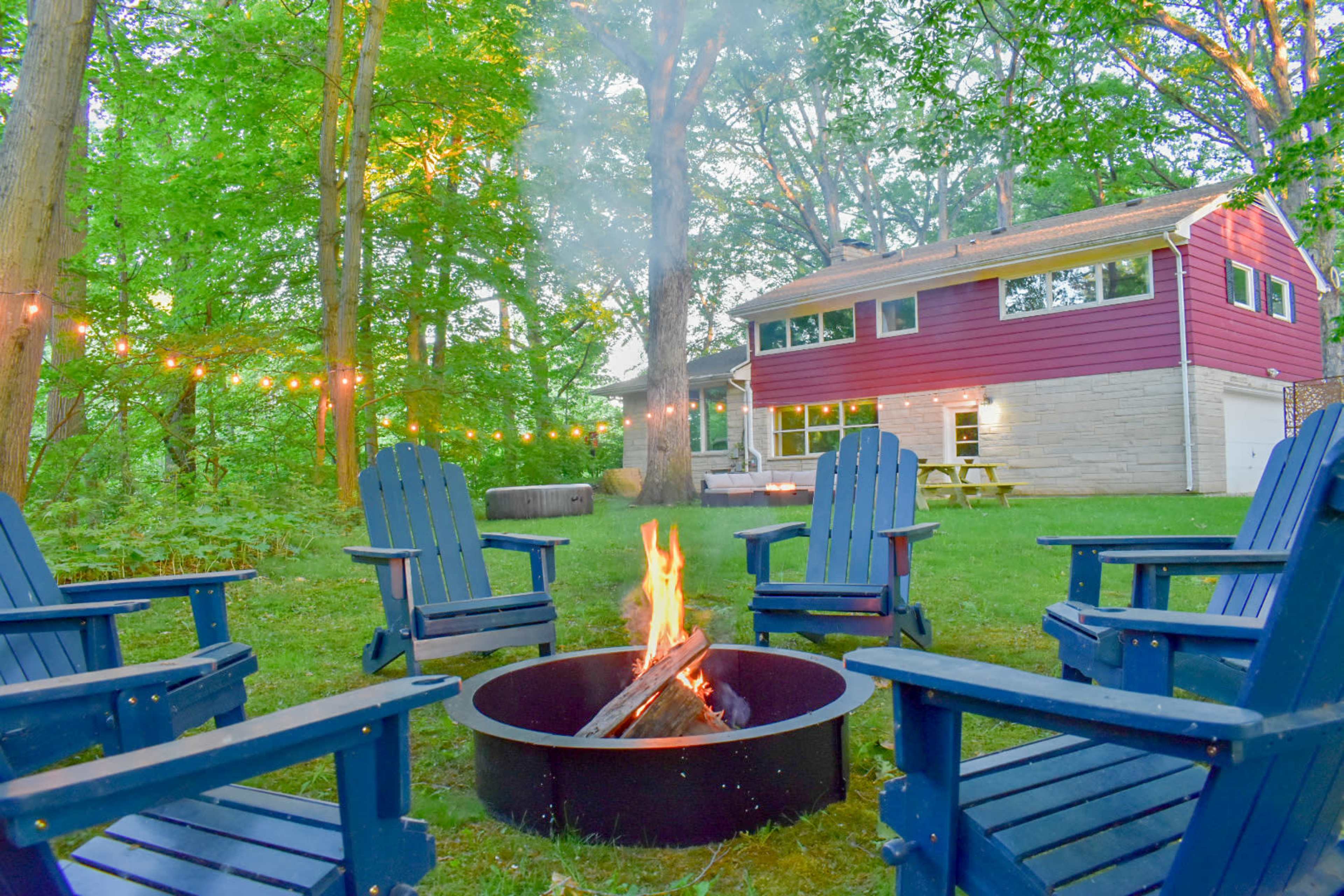 A circular fire pit surrounded by blue Adirondack chairs is located in a wooded backyard, with a house and string lights visible in the background.