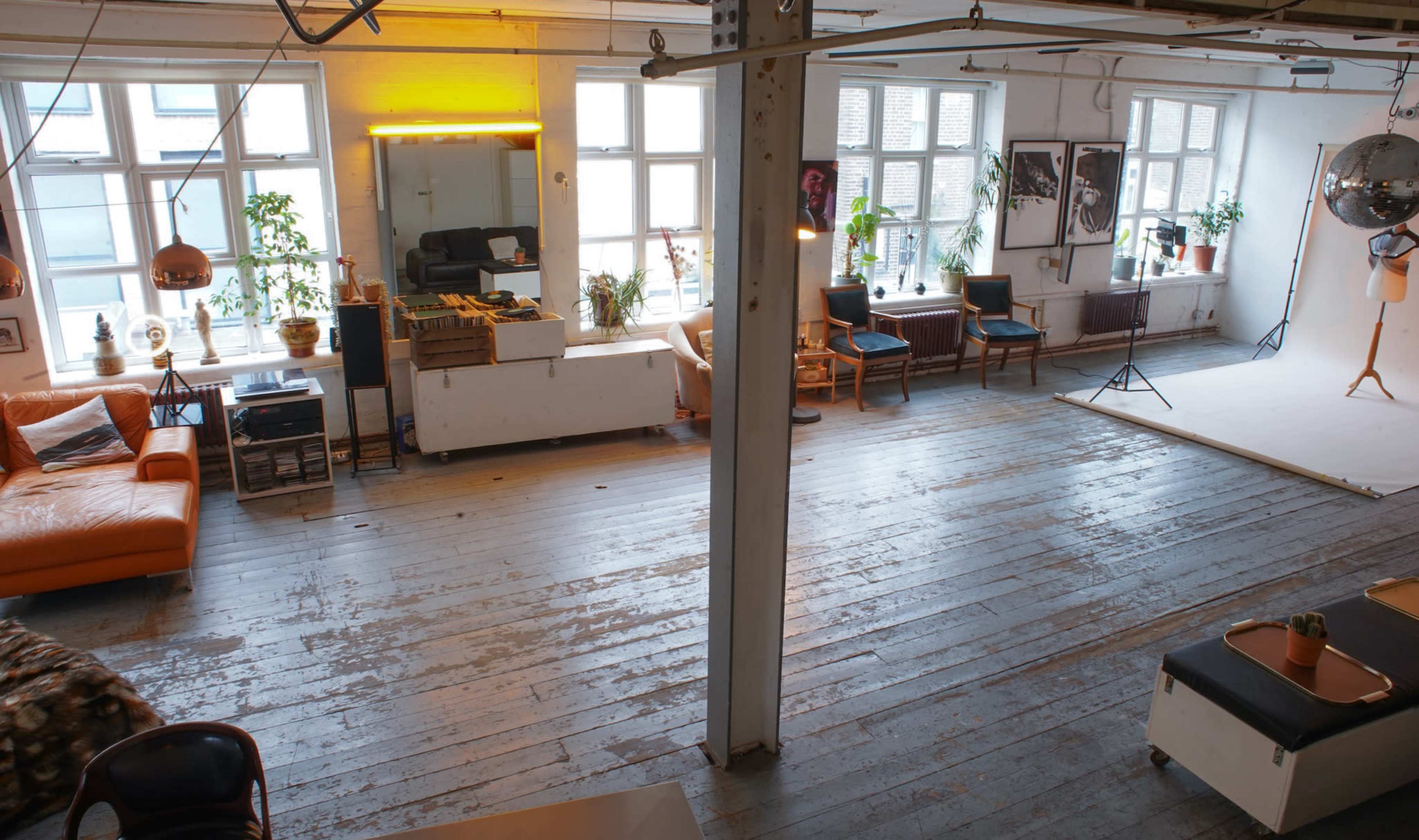 The image shows an open studio space with a wooden floor, large windows, orange sofa, and various furniture arrangements.