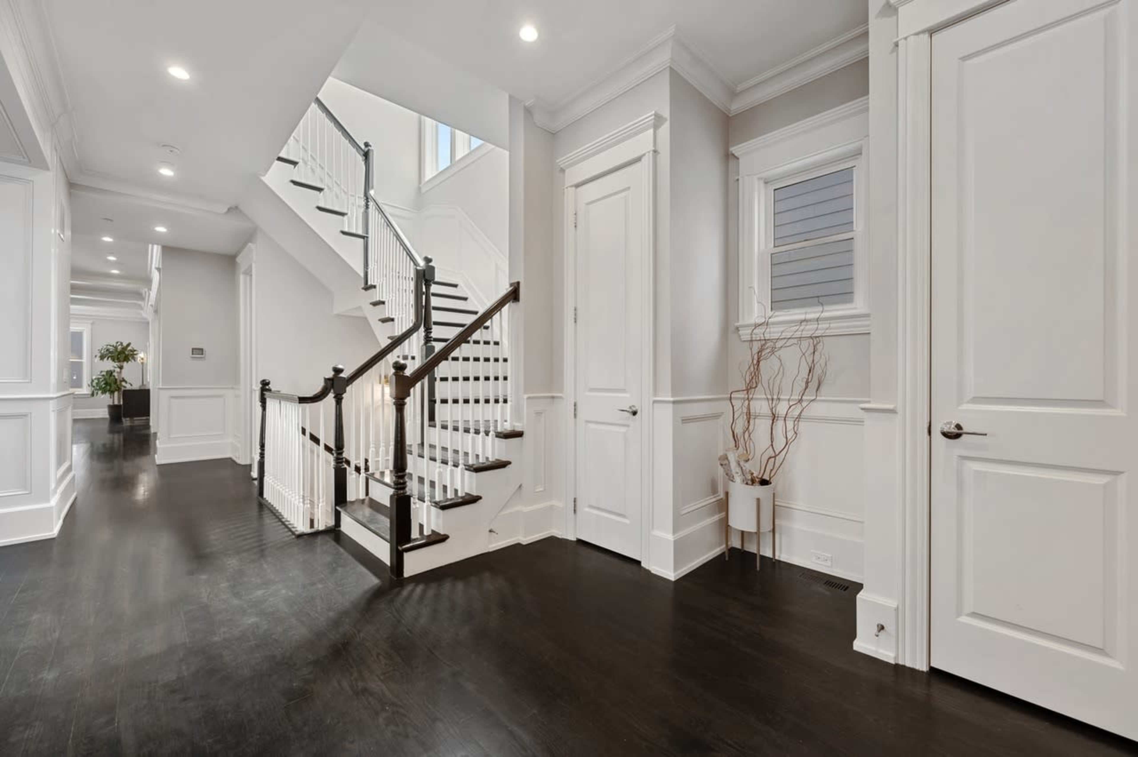 The image shows a modern foyer with a staircase and polished dark wood flooring, featuring white walls and wood accents.
