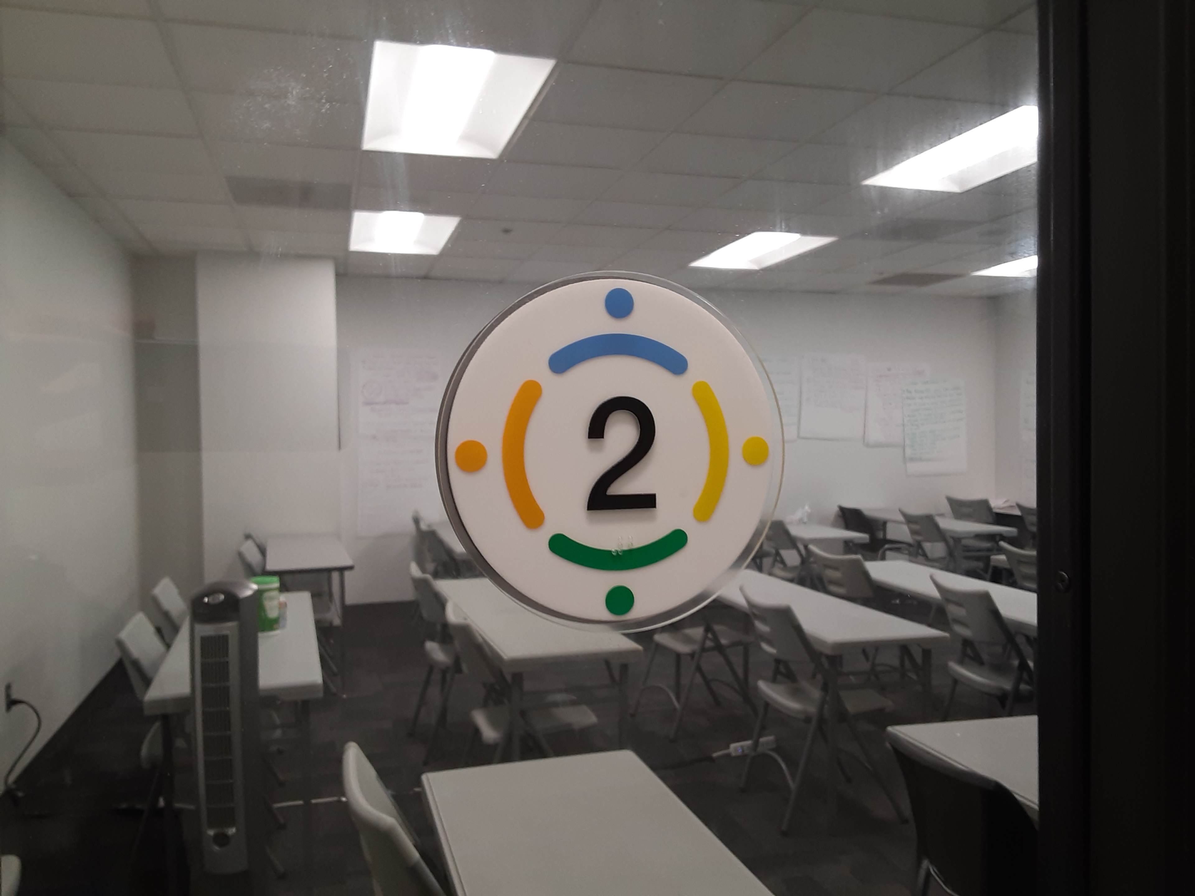 A circular sign with the number 2 and colored icons is displayed on a glass door, leading to an empty classroom with tables and chairs.