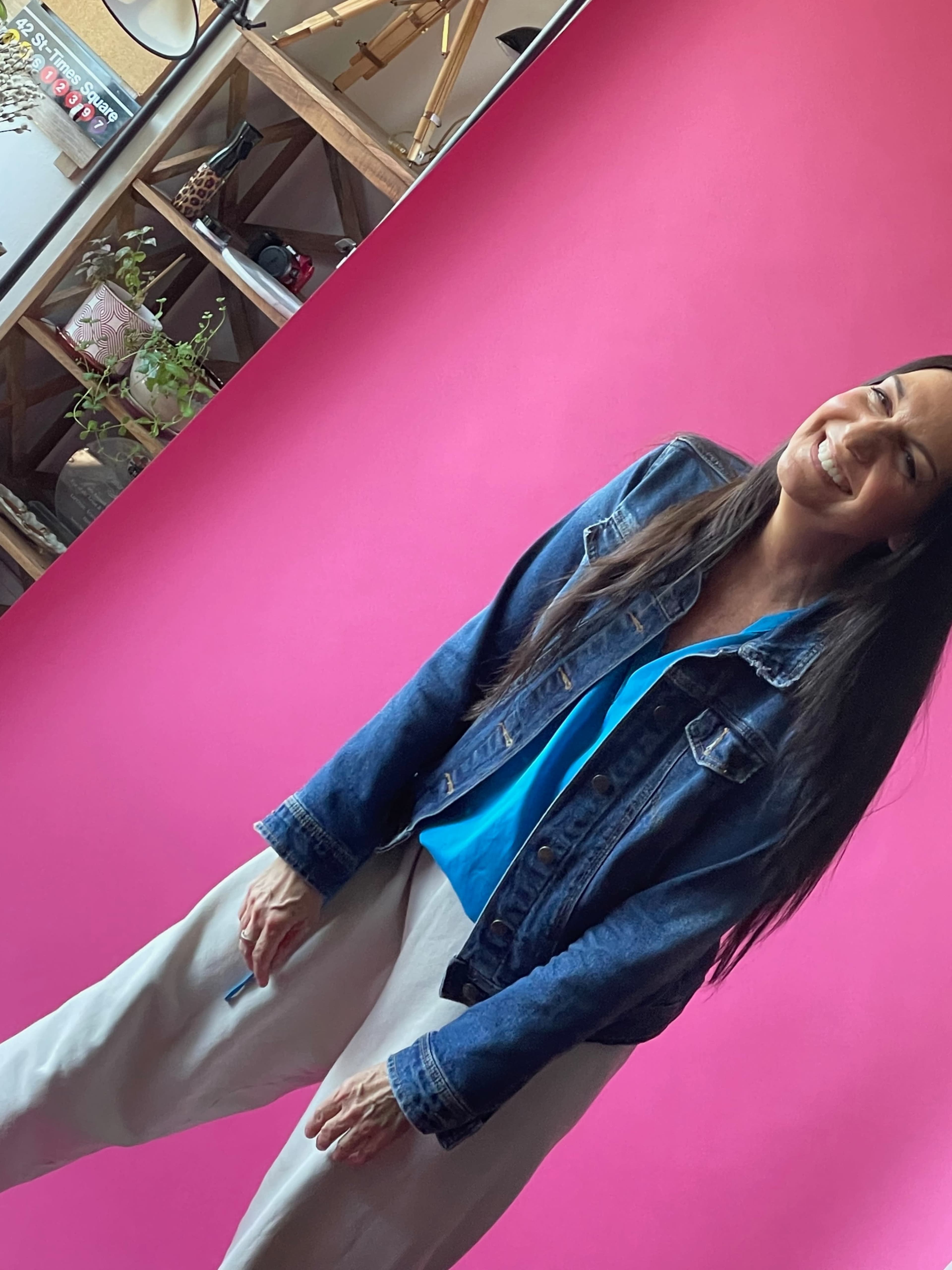 A woman in a denim jacket and blue shirt stands against a pink backdrop with a slight smile.