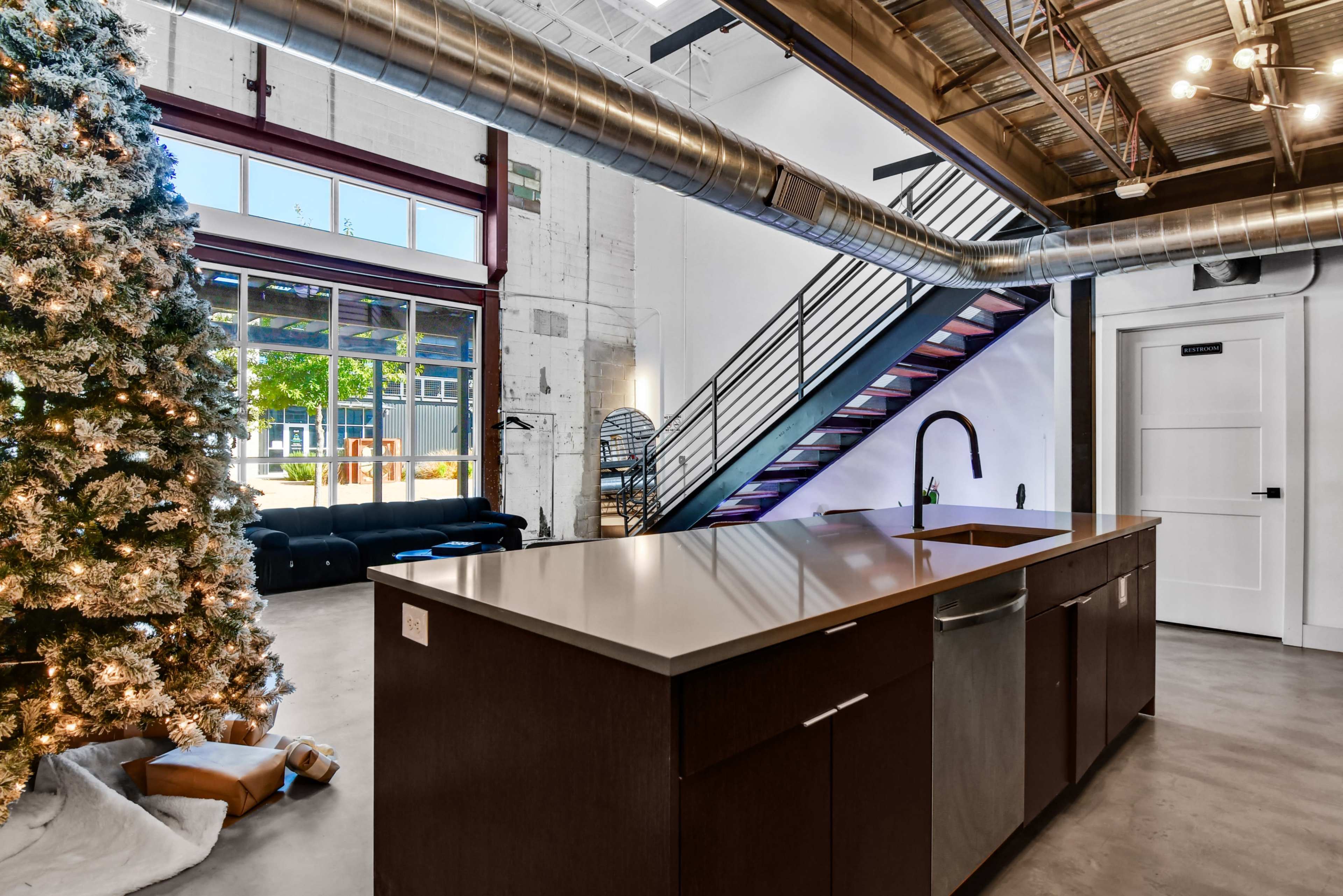 The image features a modern kitchen with a large island and a Christmas tree nearby, set in an industrial-style space with exposed beams and a staircase.