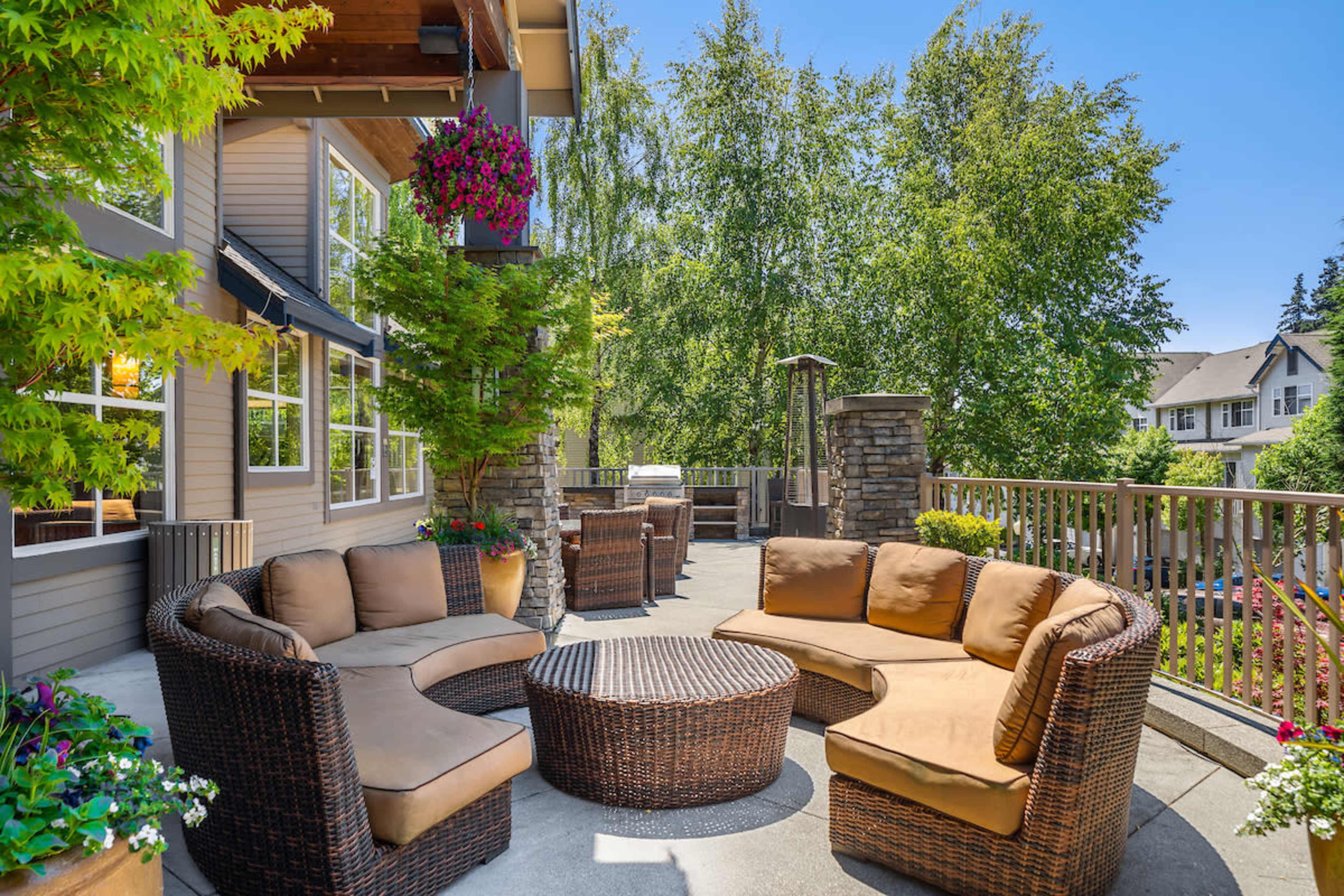 A patio features a circular seating arrangement made of wicker, surrounding a coffee table, with lush greenery and a grill in the background.