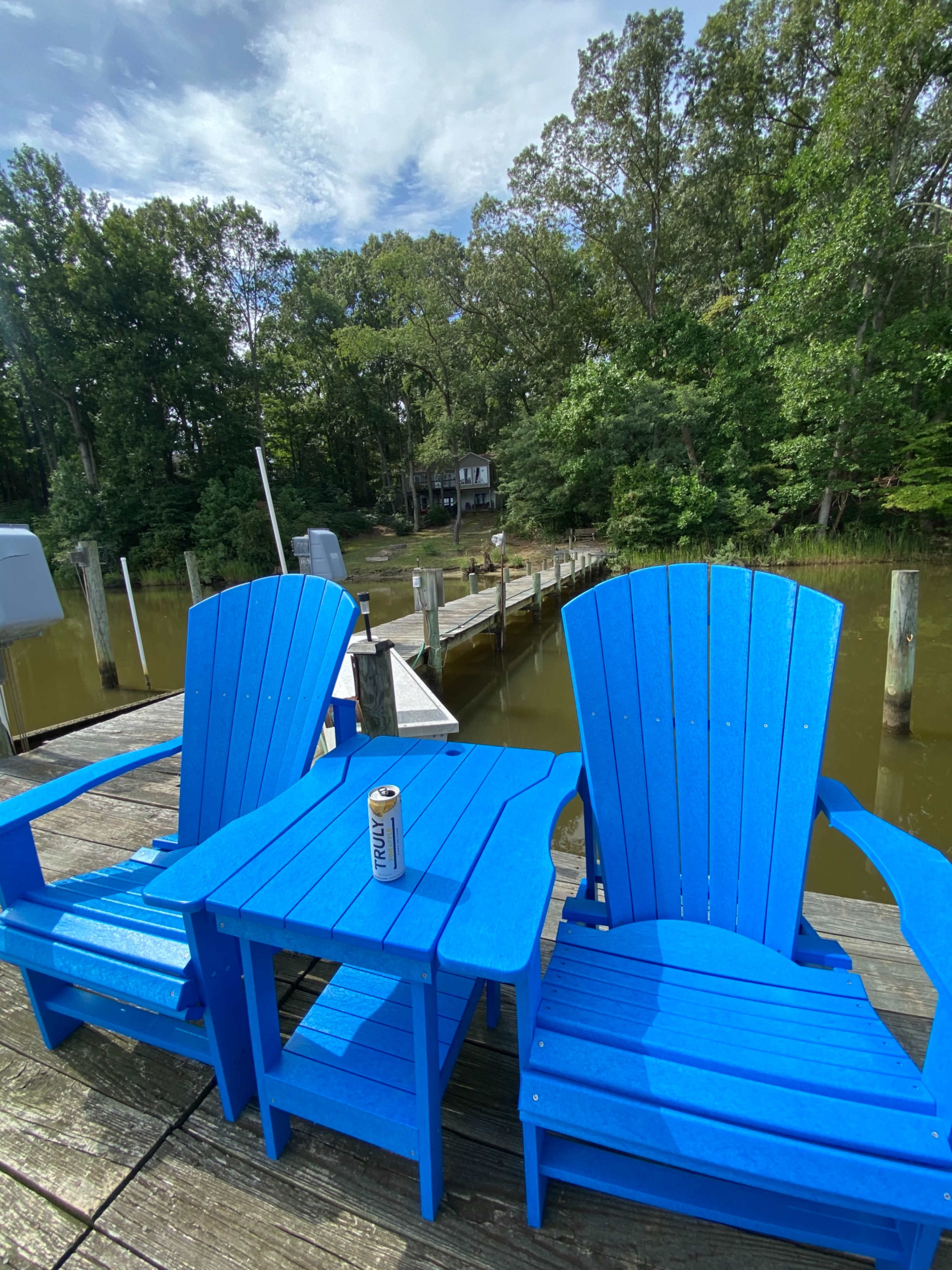 Two blue Adirondack chairs and a small table sit on a wooden deck overlooking a calm waterway lined with trees.