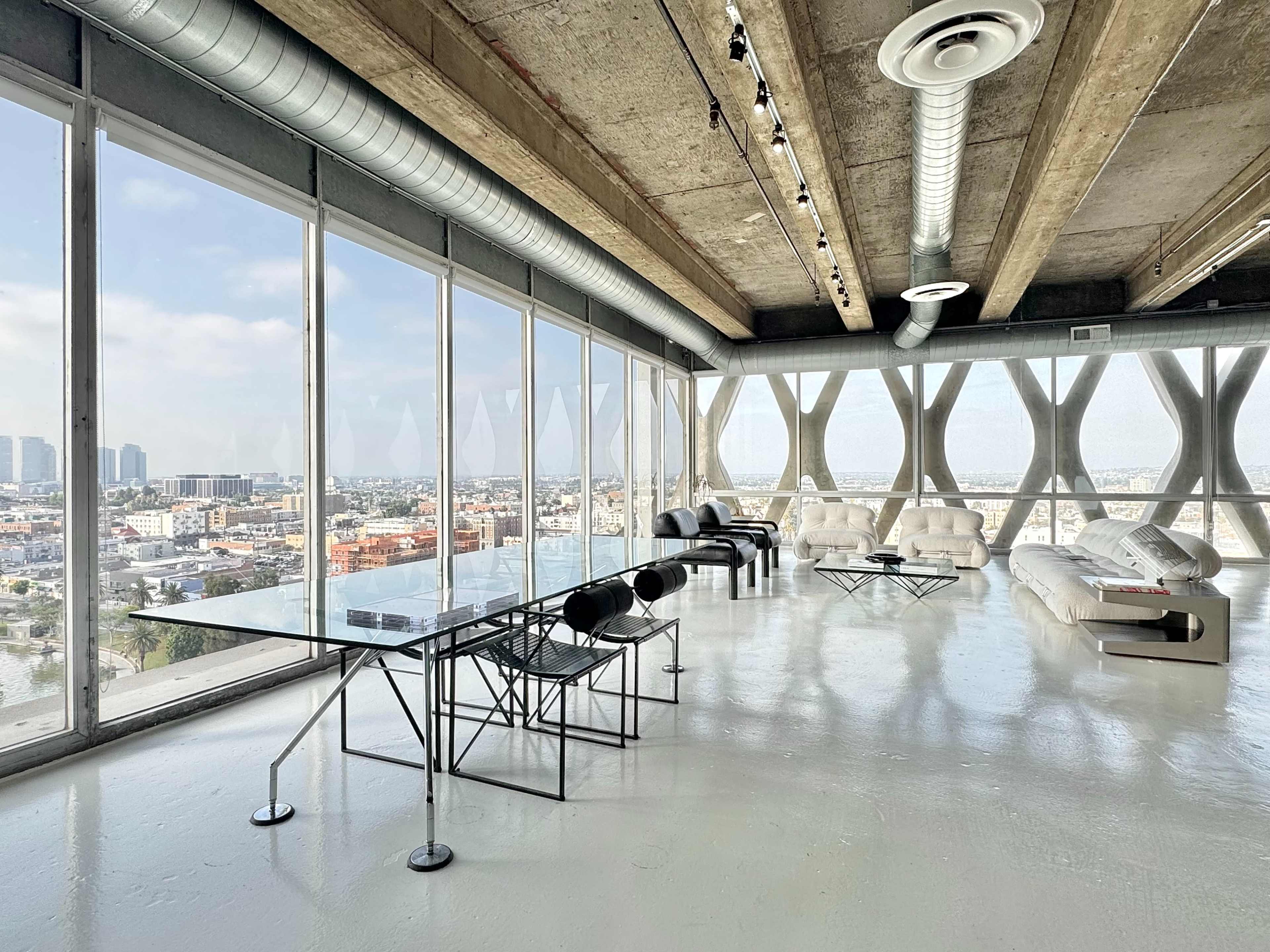 The image shows a spacious, modern room with large windows offering a panoramic view of a city skyline, featuring minimalistic furniture including a glass dining table and white sofas.