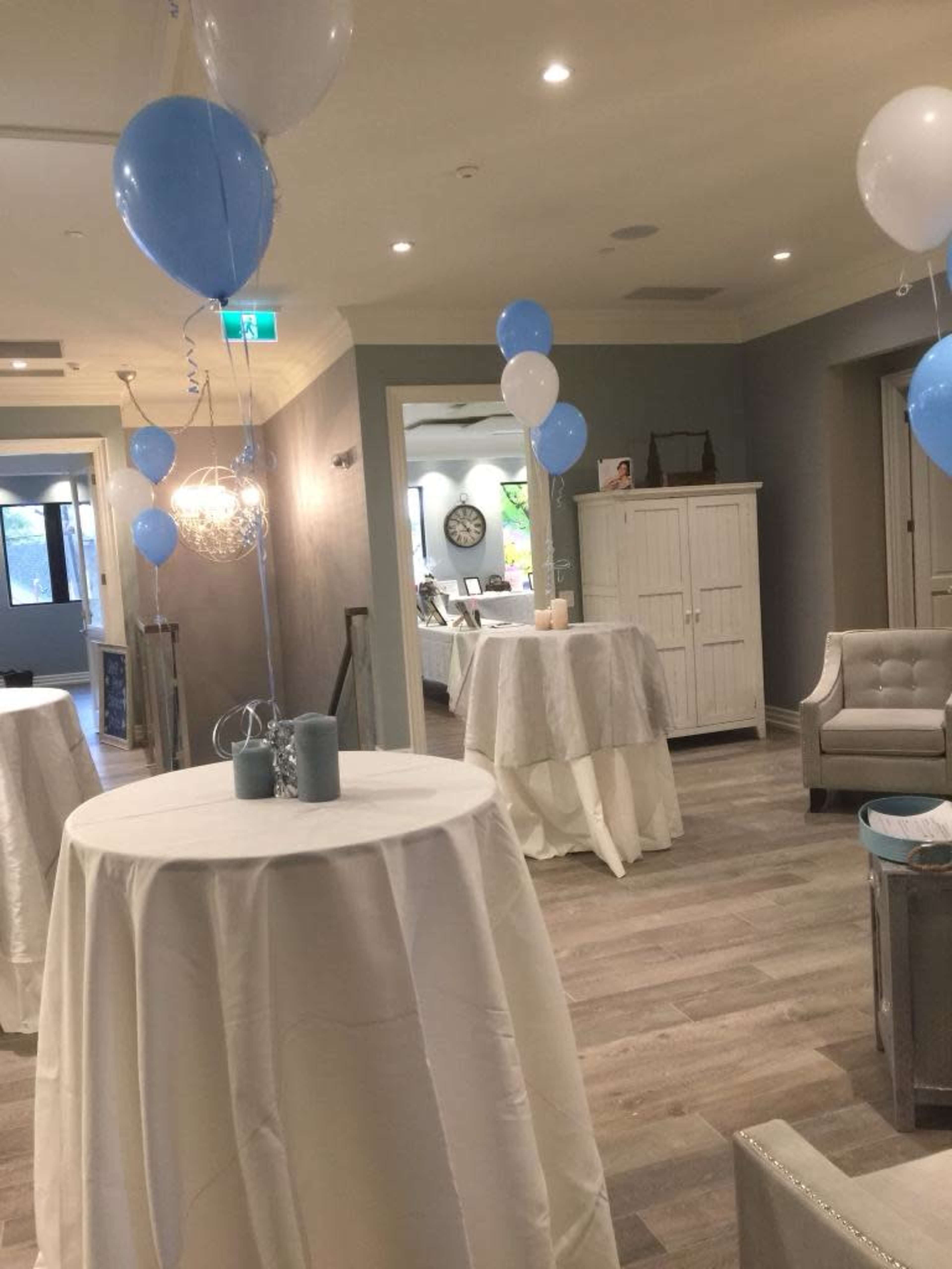 A decorated indoor space with tables covered in white tablecloths, blue and white balloons, and soft lighting.