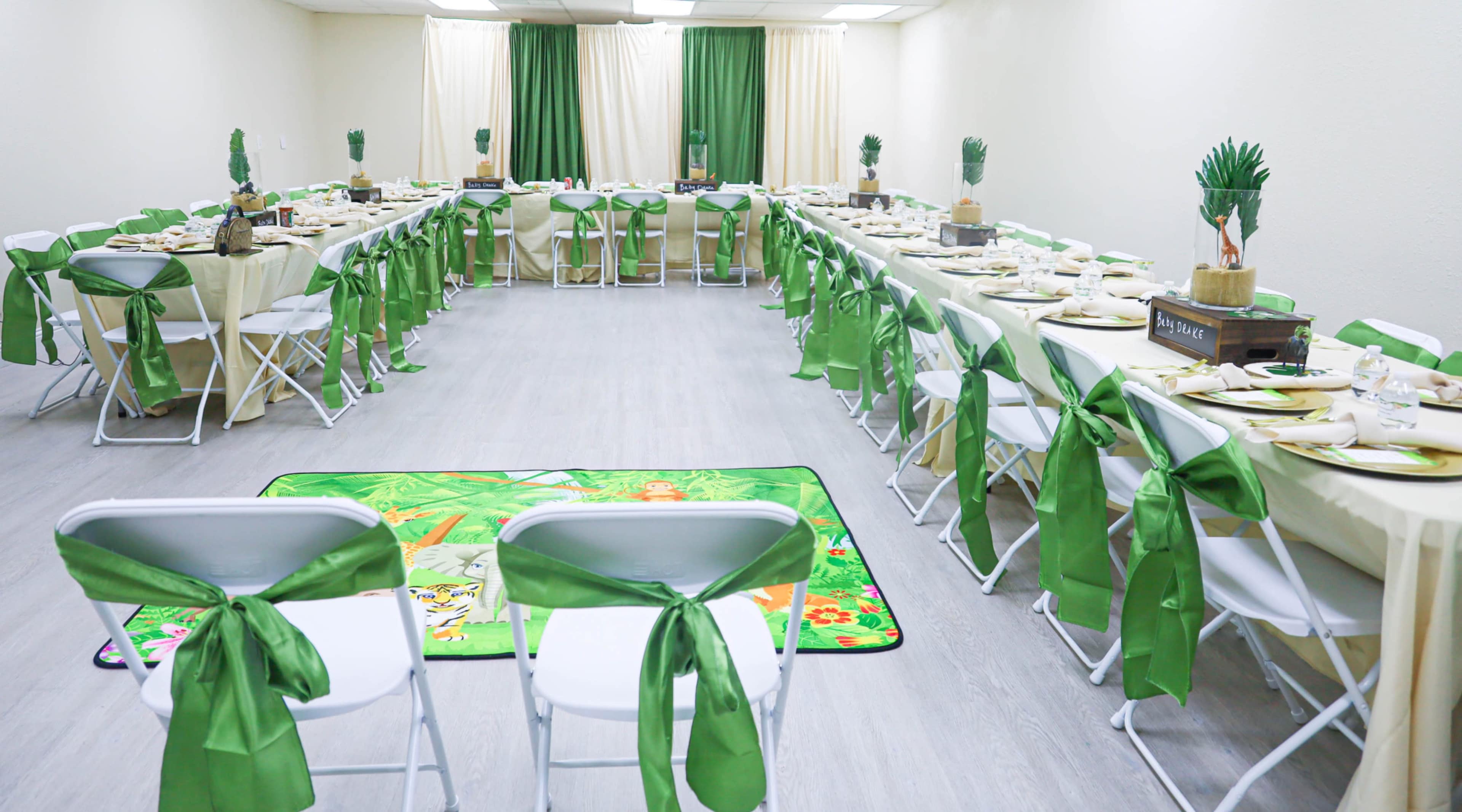 A banquet hall set up for an event with two long tables covered in cream and green tablecloths, arranged with dishes and decorations, and surrounded by white chairs adorned with green ribbons.