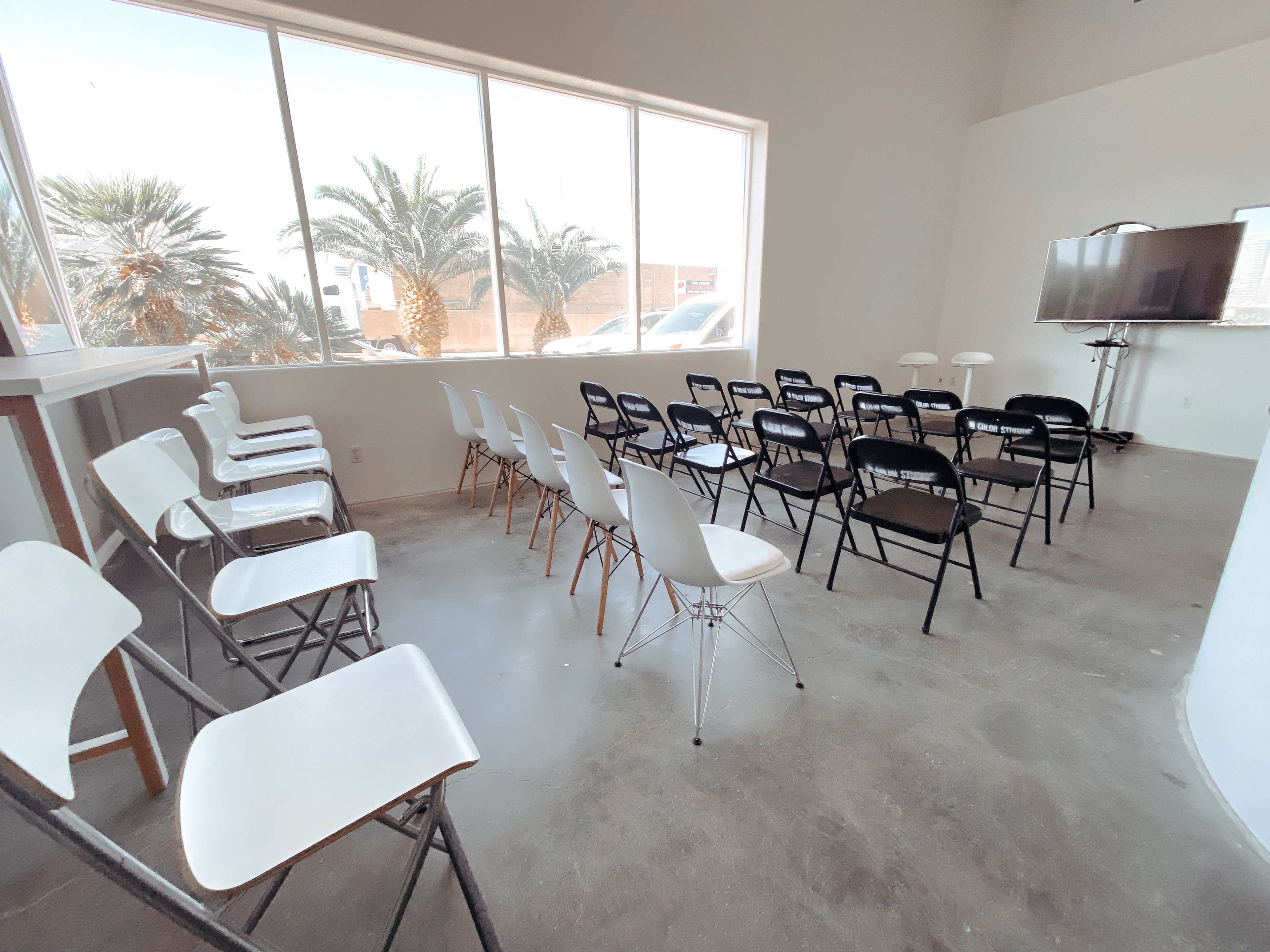 A meeting room features rows of black and white chairs arranged facing a television on a stand, with large windows showcasing palm trees outside.
