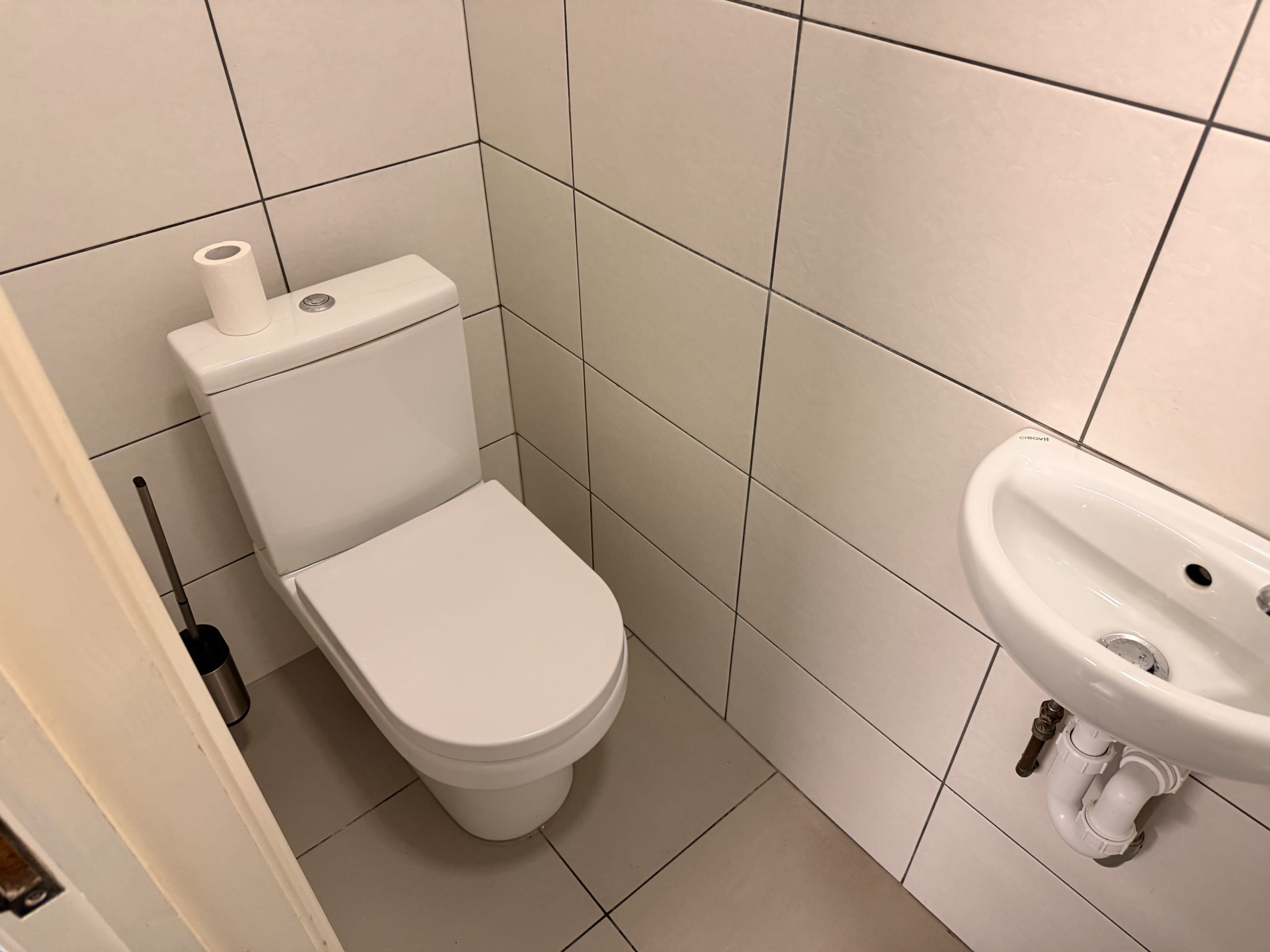 The image shows a small restroom with a white toilet and a sink against tiled walls.
