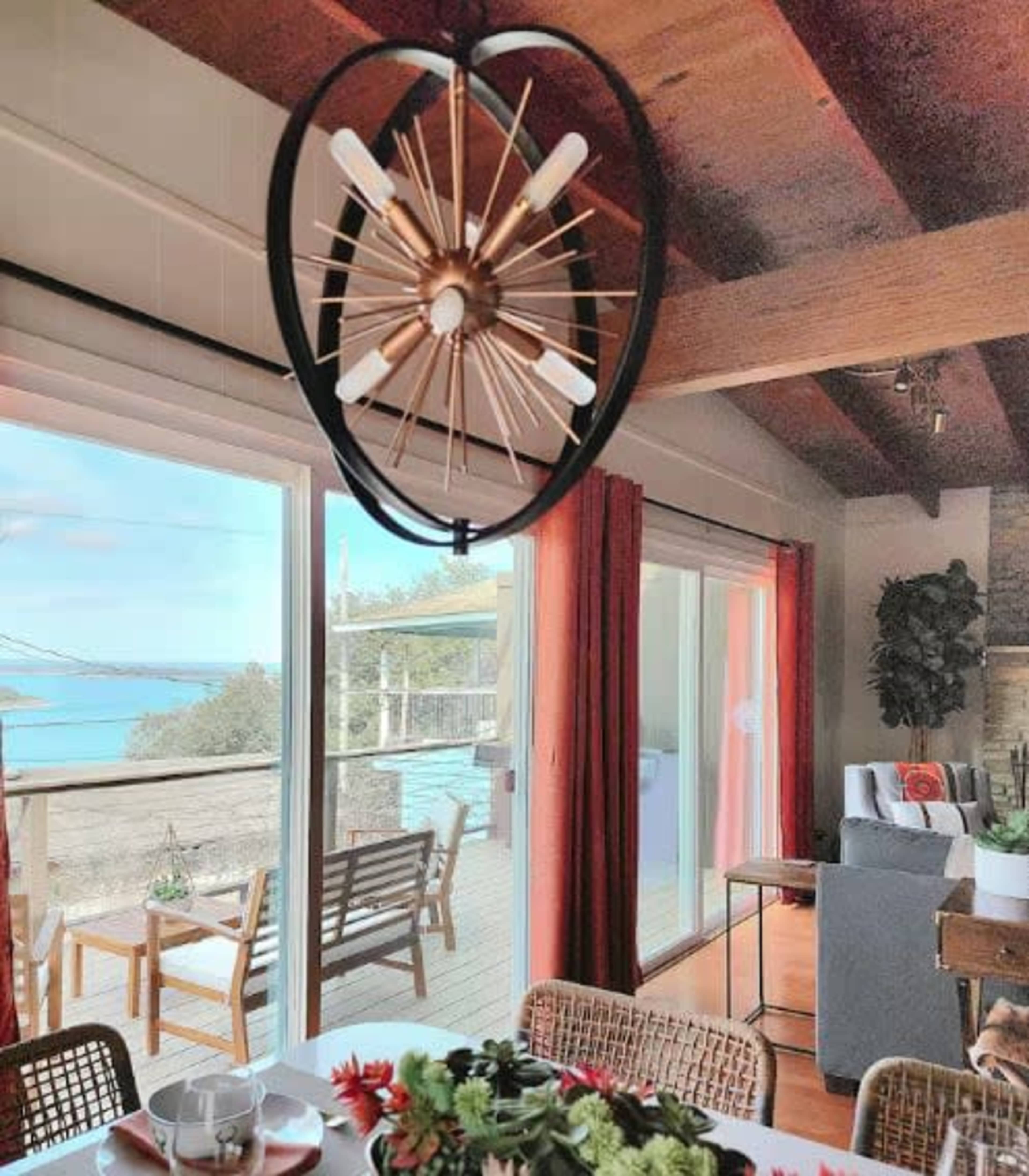 A modern dining area features a light fixture hanging above the table with a view of a deck and ocean beyond large windows.