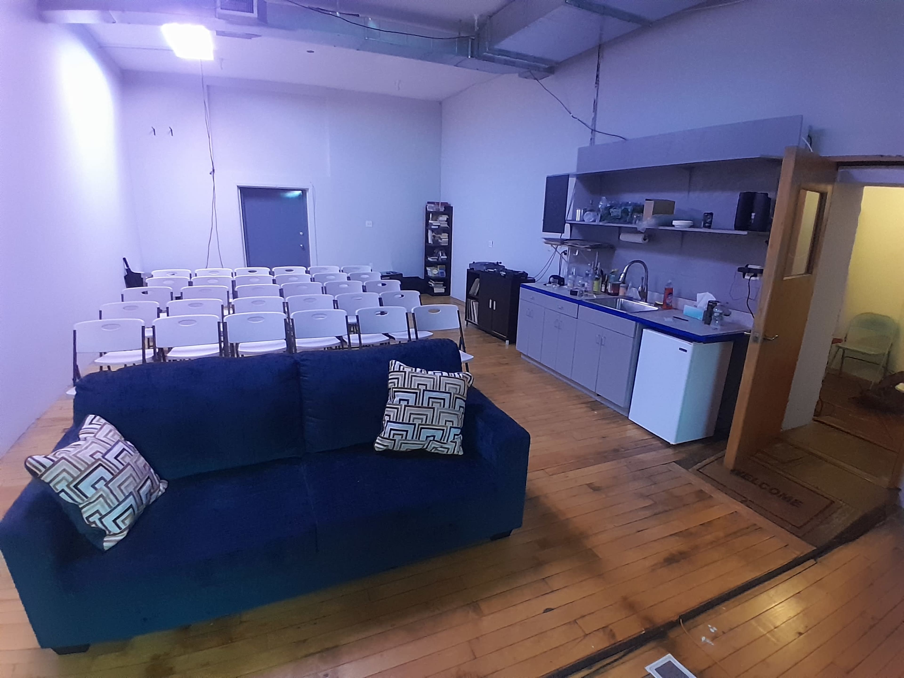 The image shows a sparsely furnished room with a blue couch, a kitchenette area, a set of chairs arranged for a meeting, and a doorway leading to another space.