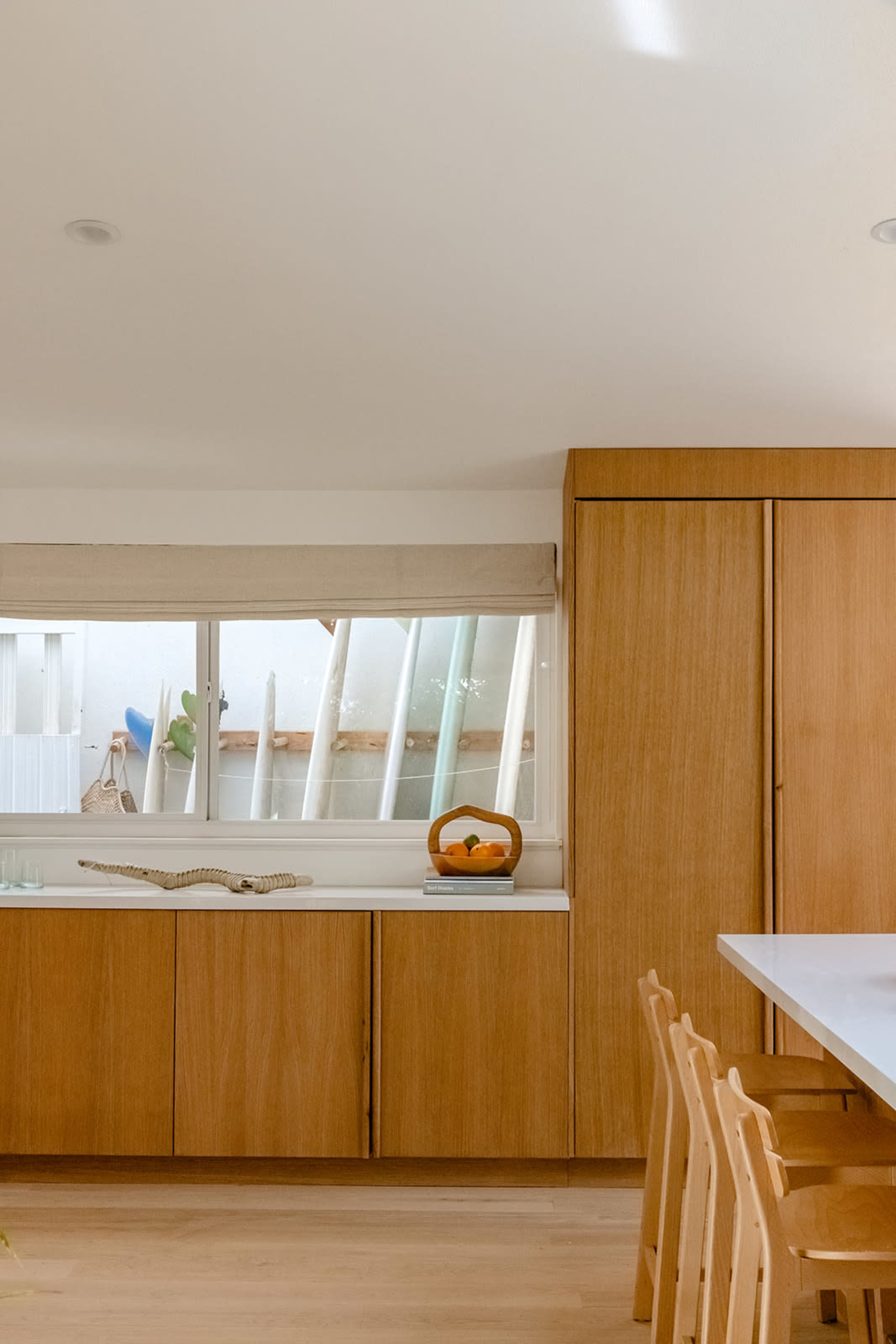 A modern kitchen features wooden cabinetry, a large window with a subtle shade, and a countertop with a fruit basket.