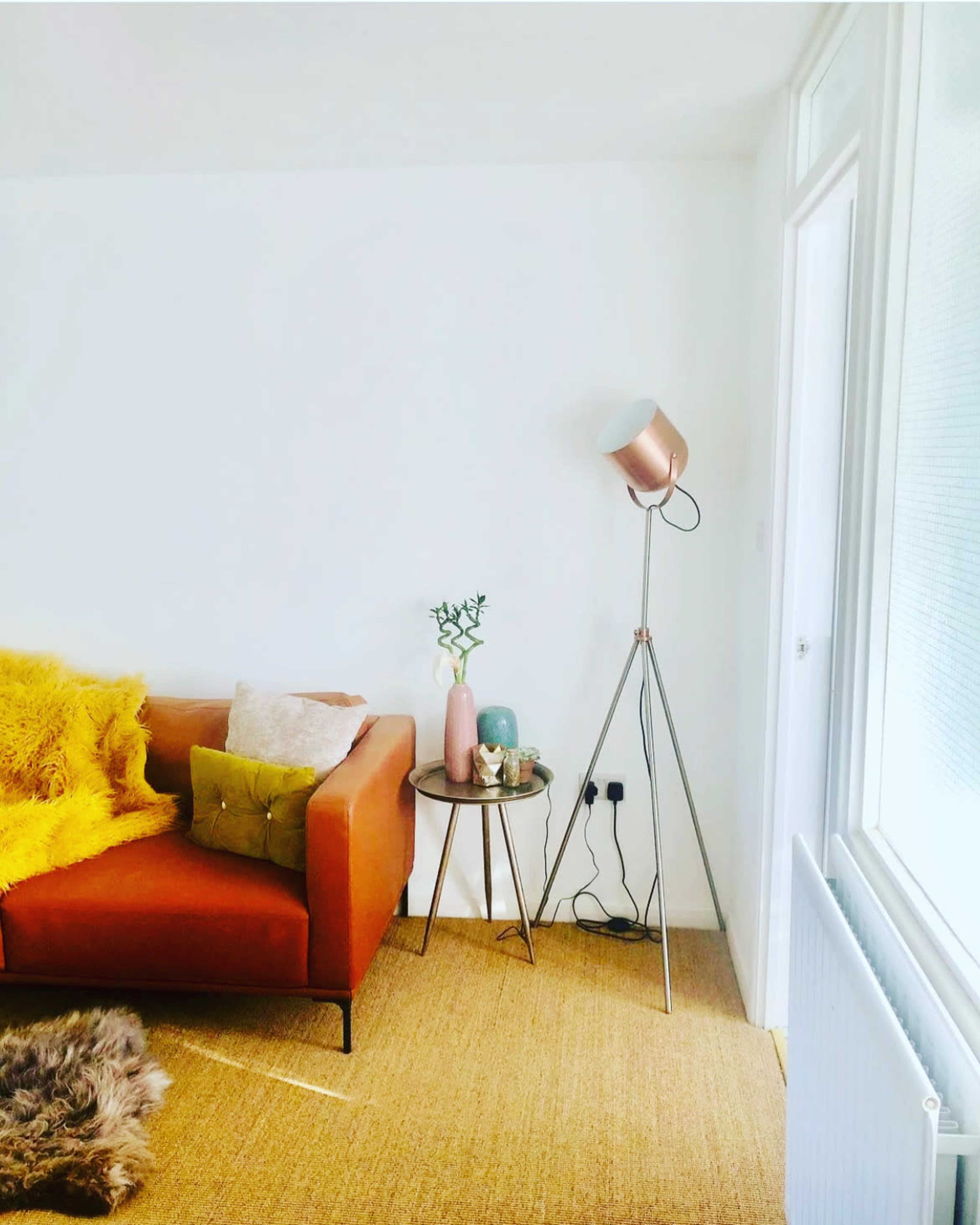 The image shows a cozy living room featuring an orange sofa with decorative cushions, a round side table with vases, a modern floor lamp, and a textured rug on a natural fiber floor.