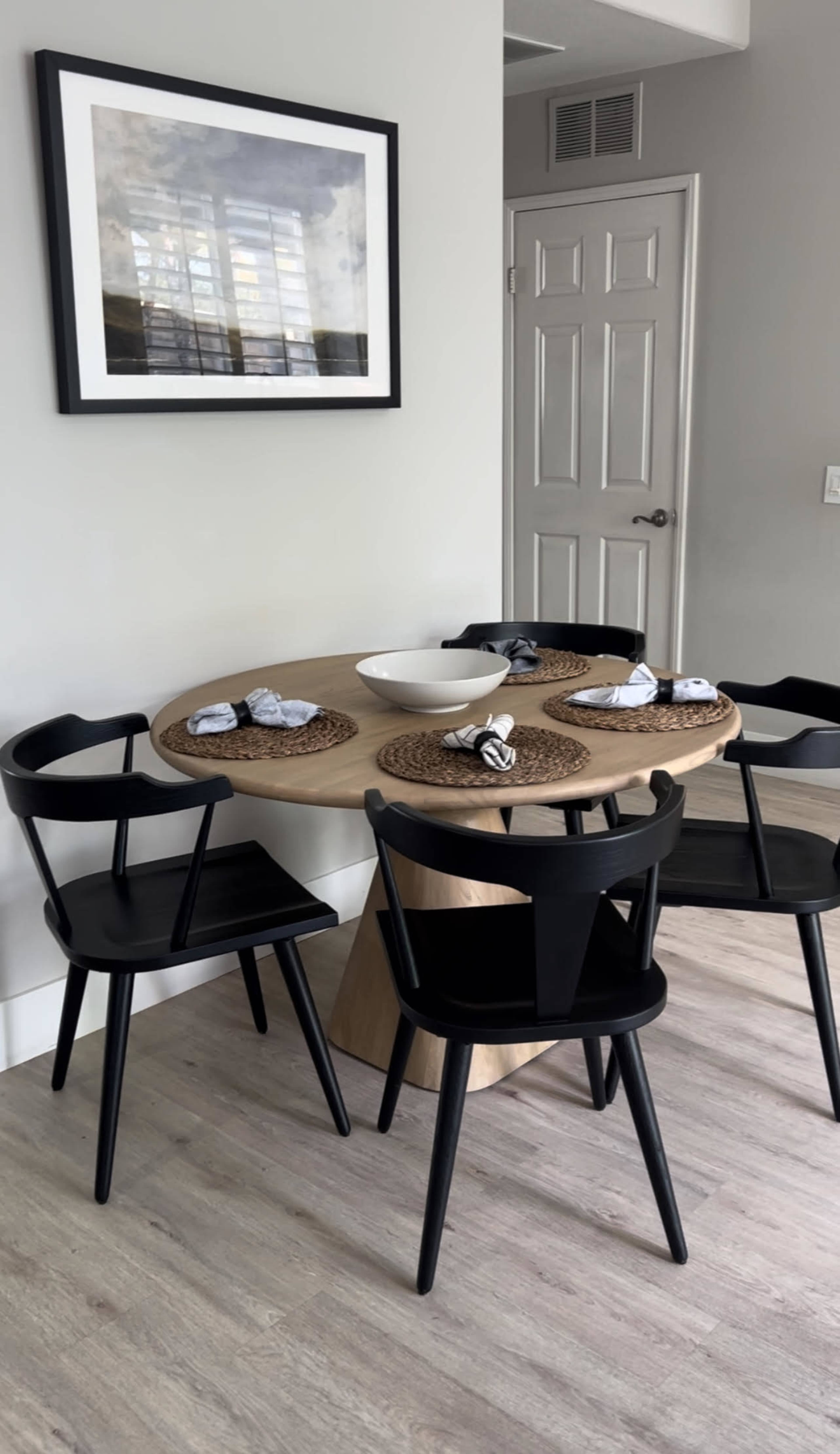 A round wooden dining table is set with four black chairs and decorative place settings, situated against a light-colored wall with a framed artwork above.