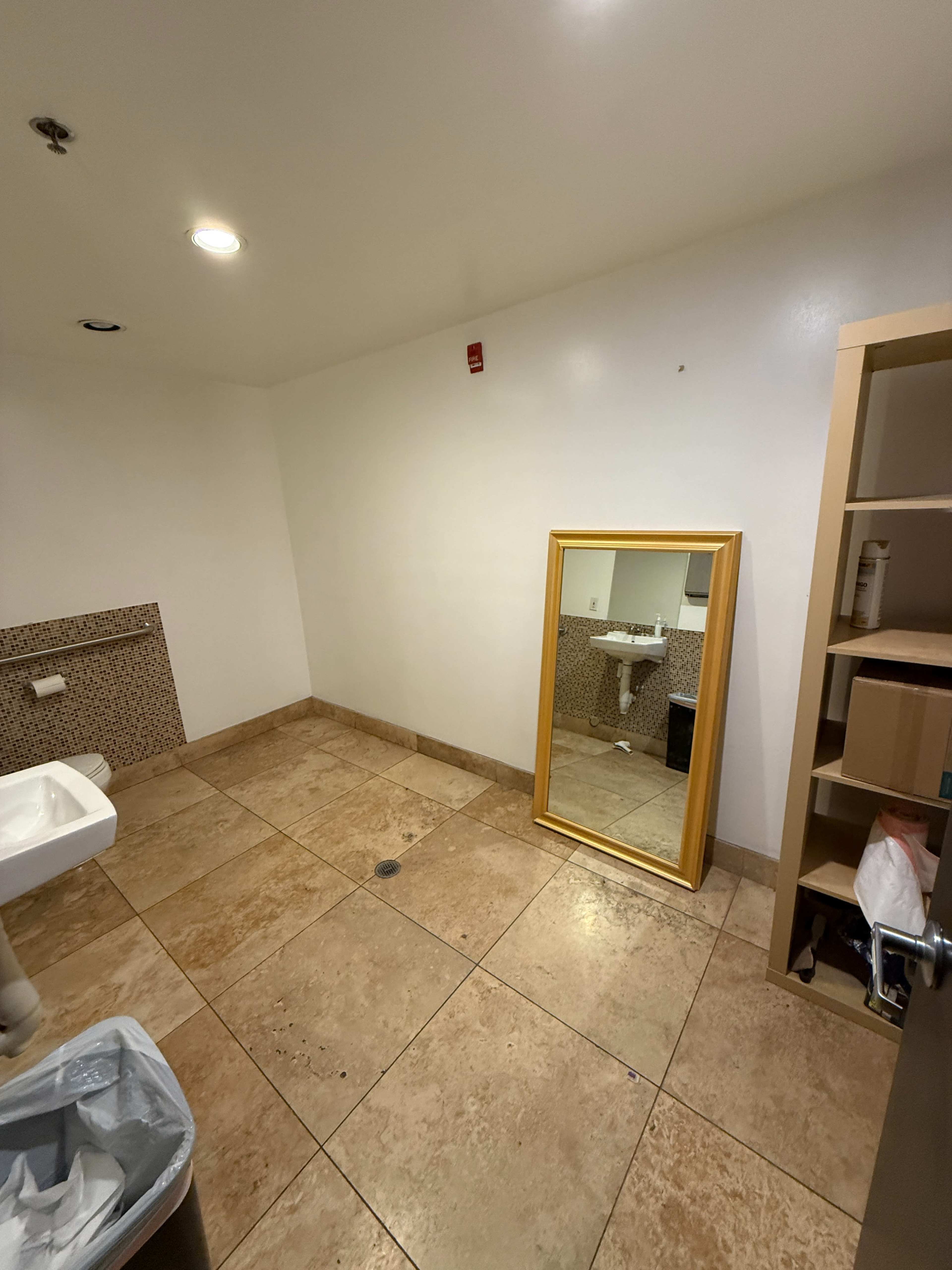 The image shows an empty bathroom with tiled floors, a wall-mounted sink, a large mirror leaning against the wall, and a trash can in the corner.