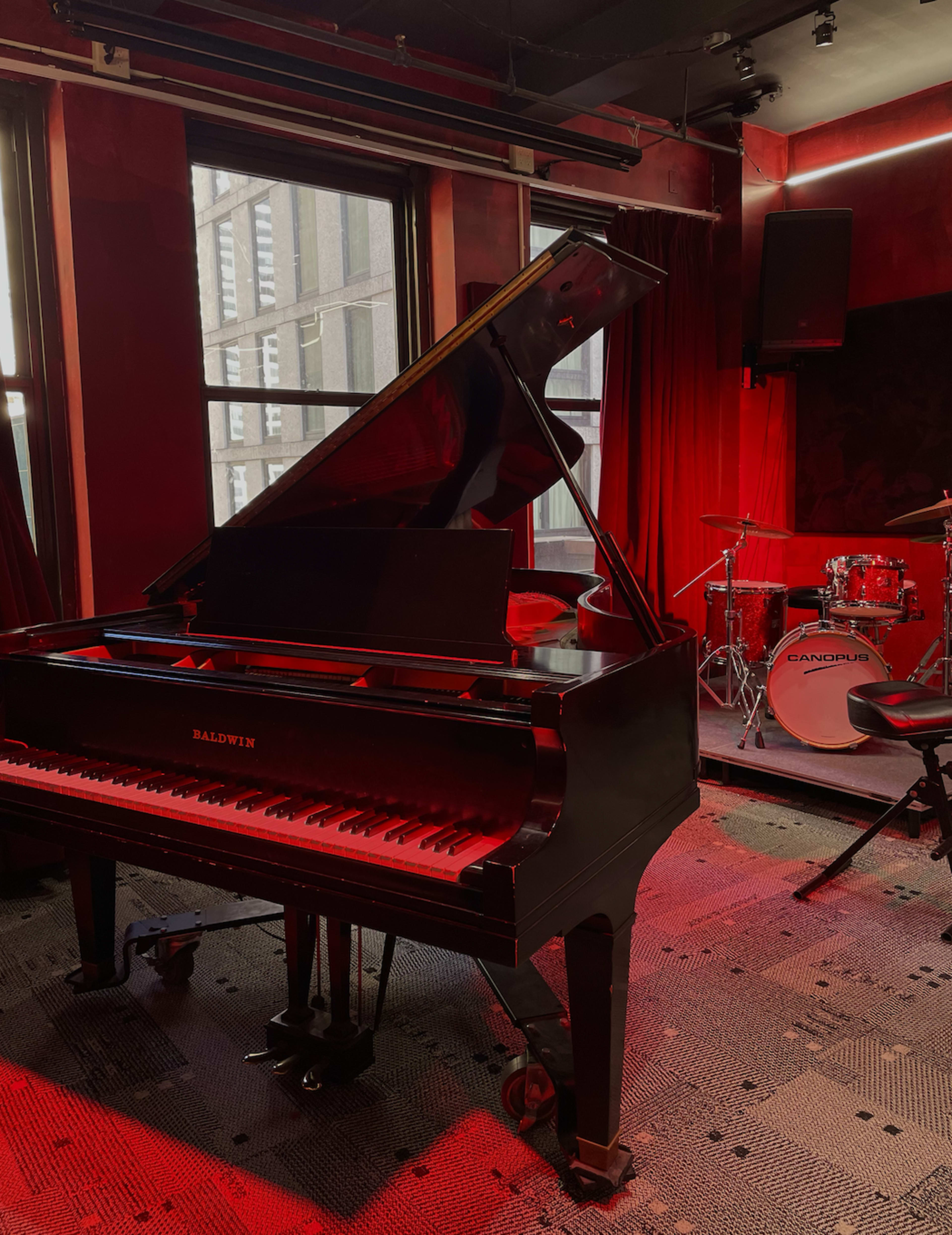 A grand piano sits in a dimly lit room with red curtains, next to a small drum set.