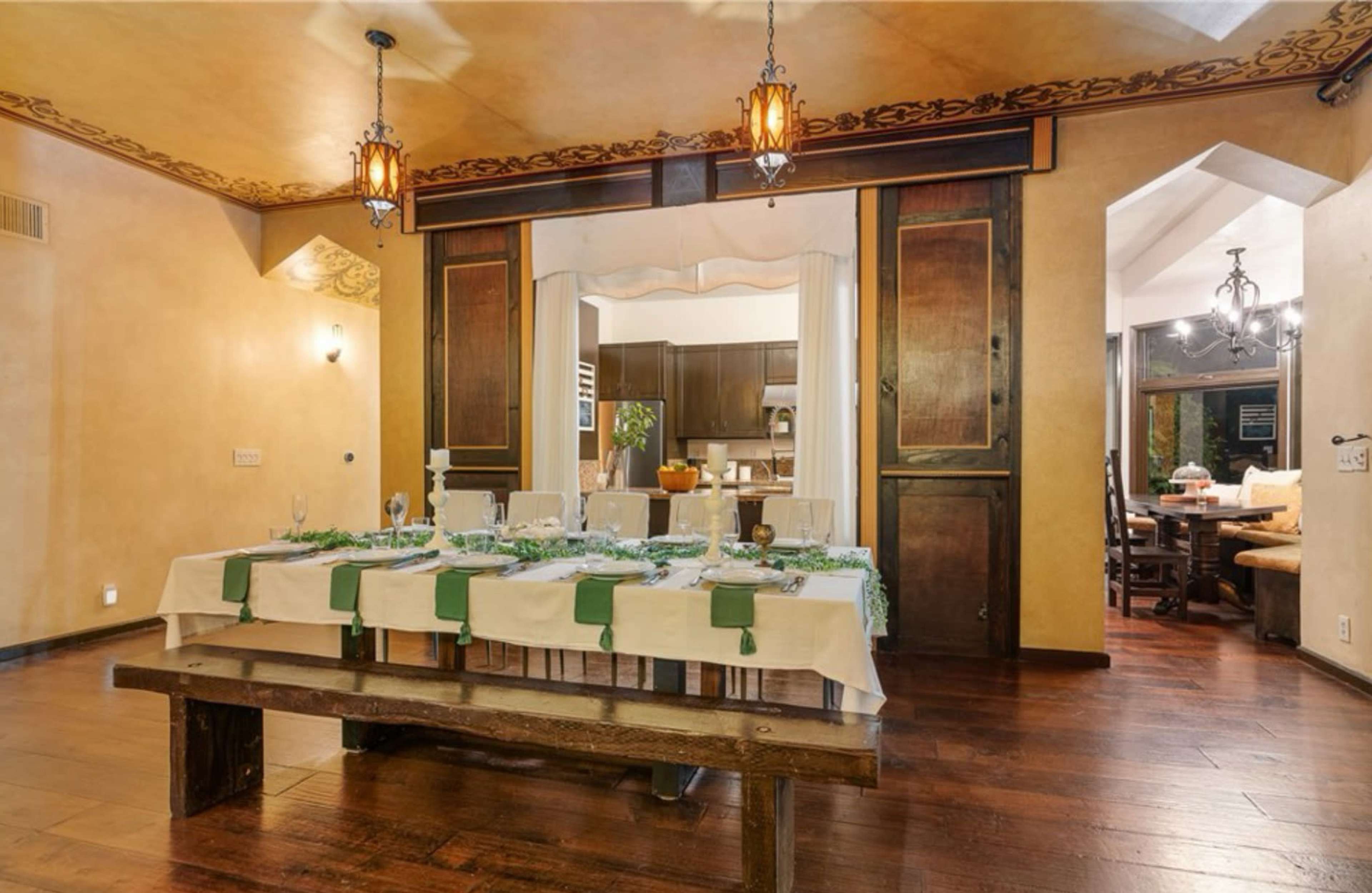 A long wooden dining table is set with white tablecloths and green accents in a warmly lit dining area with wooden beams and decorative lanterns.