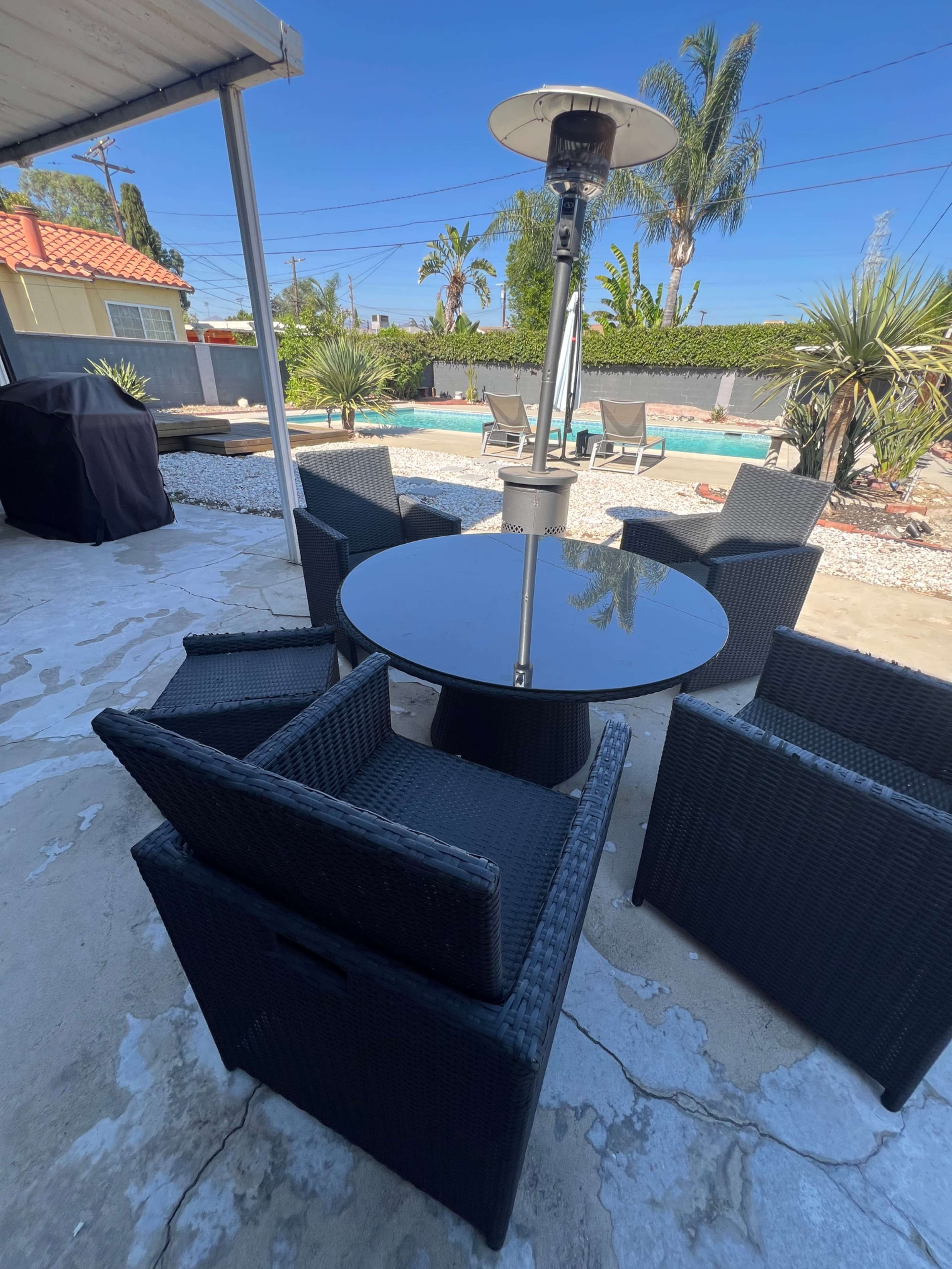 A black round table surrounded by four wicker chairs is situated on a patio near a backyard pool.