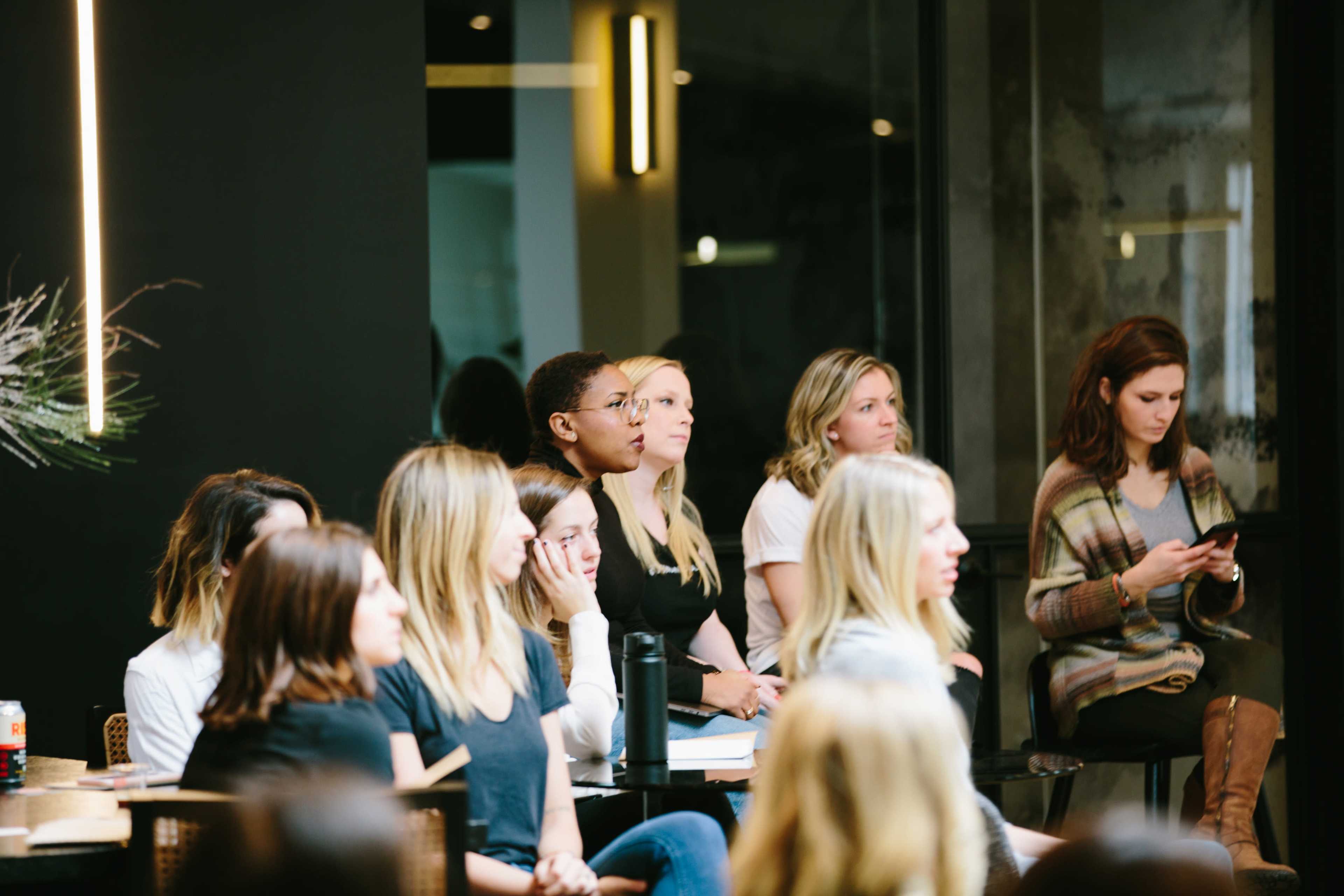 A group of women sits in a modern space, attentively listening to a speaker.