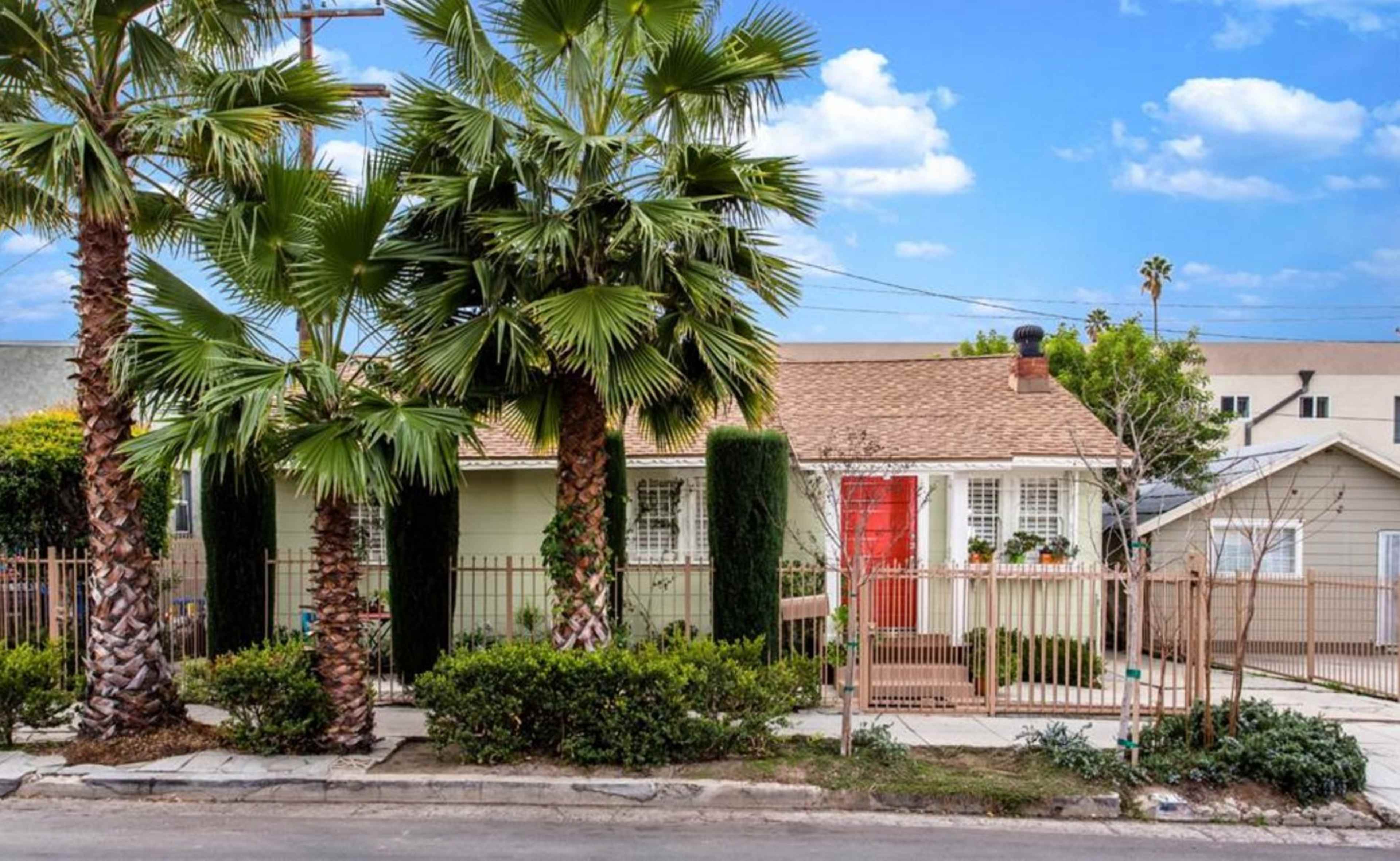 A single-story house with a red door, surrounded by palm trees and a low fence, located on a residential street.