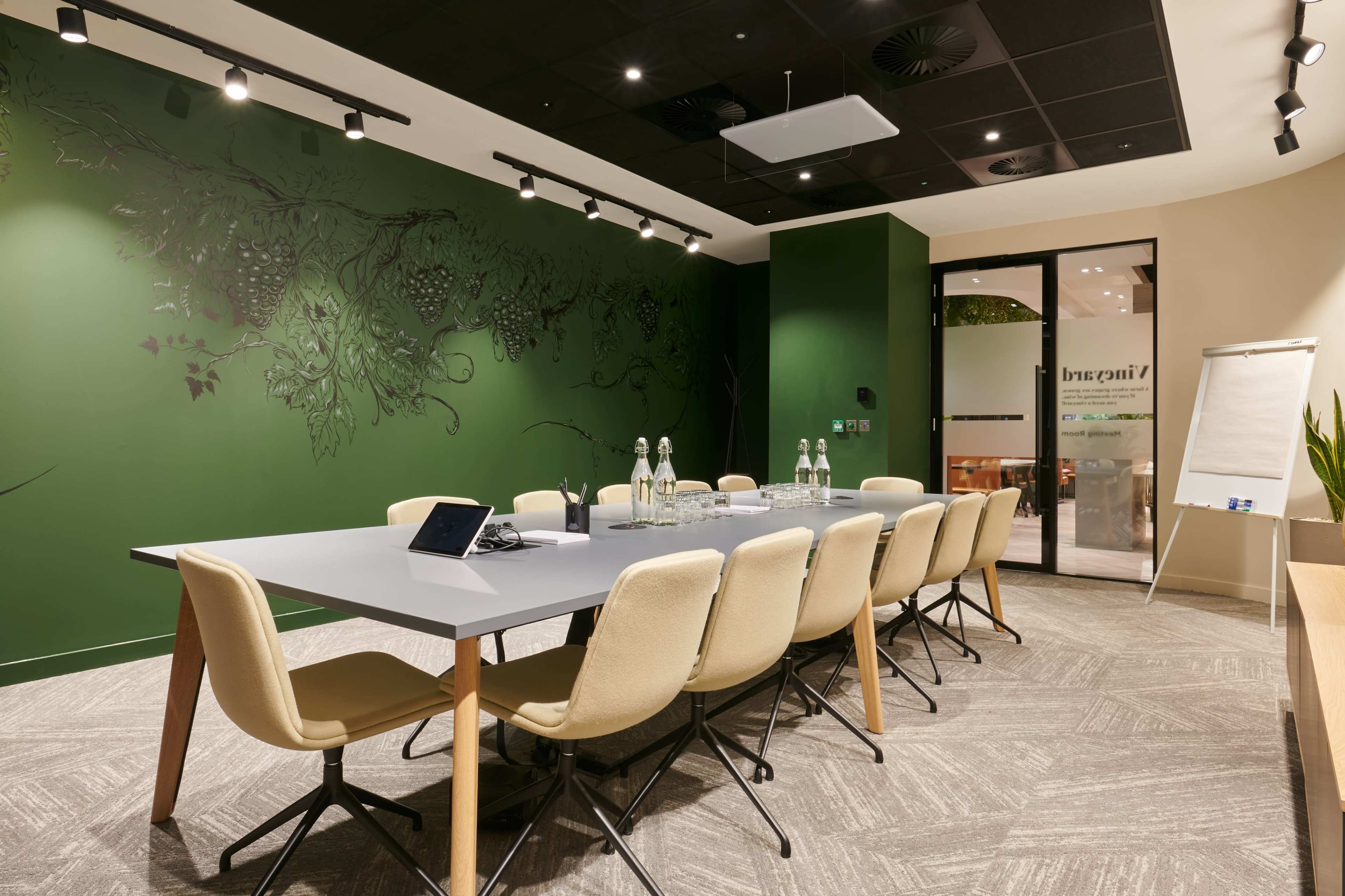 A modern conference room features a long table with beige chairs, green walls adorned with a vine mural, and large windows allowing natural light.