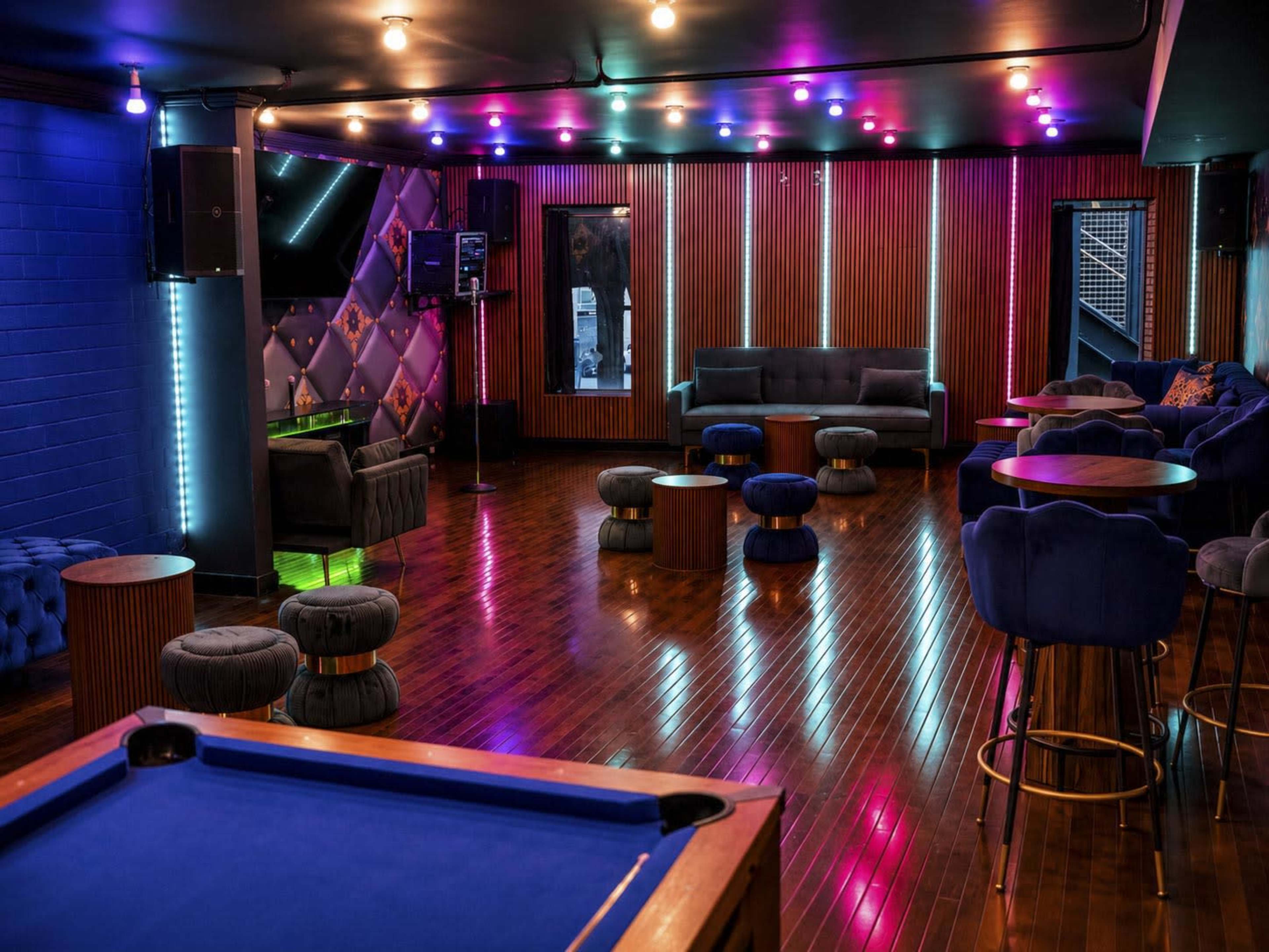 The image shows a modern lounge space with colorful lighting, plush seating, and a pool table in the foreground.