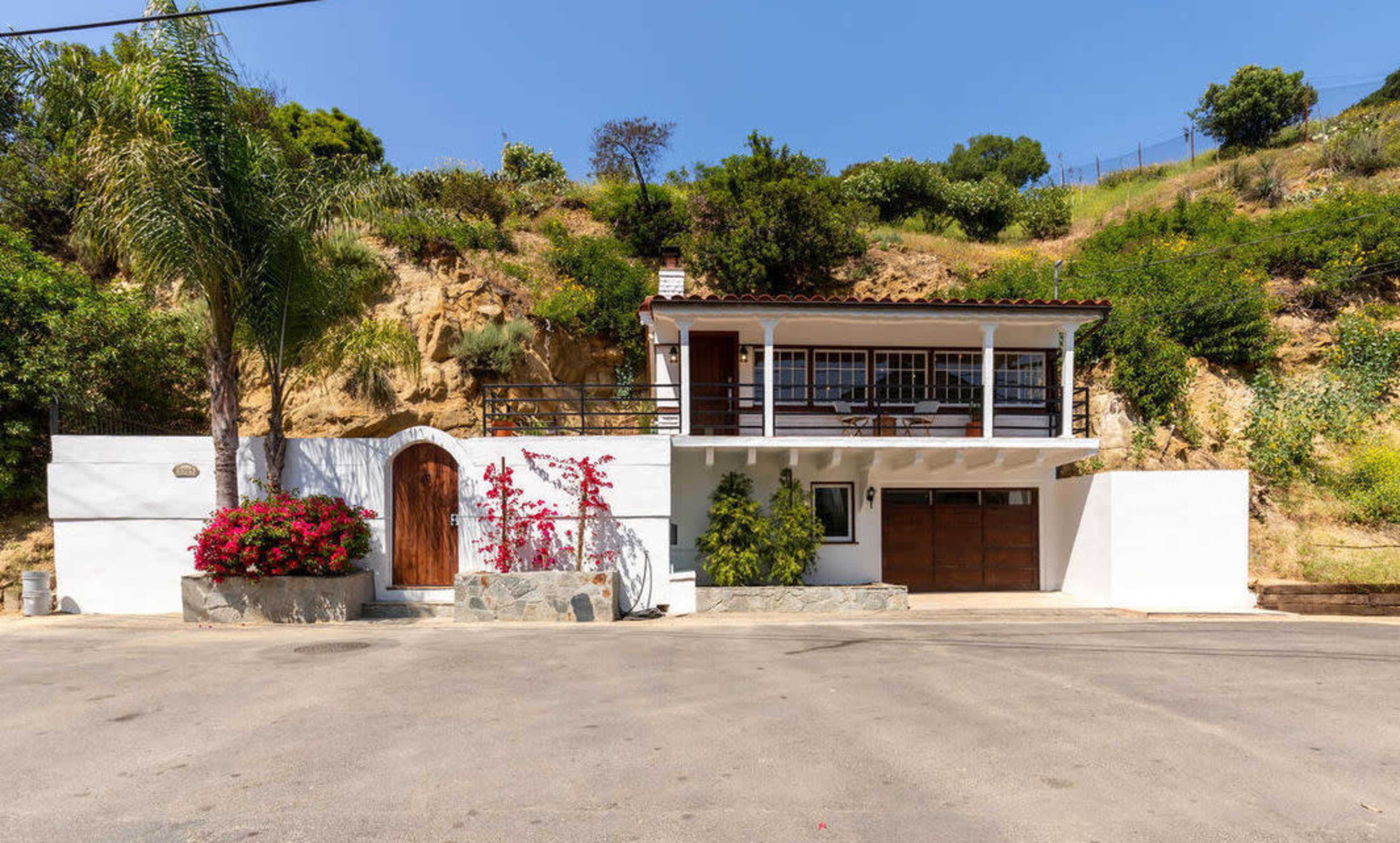 The image shows a two-story house situated on a sloped lot, with a driveway and a garden featuring colorful flowers in front.