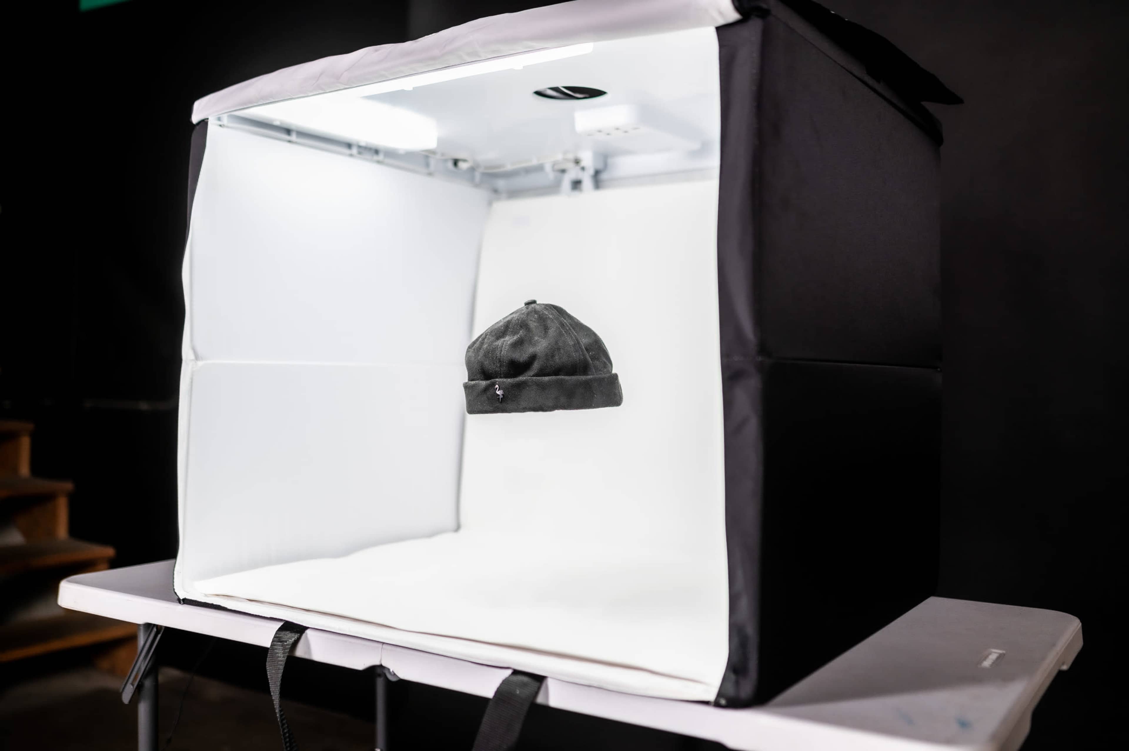 A black hat is suspended inside a lighted photography tent on a table.