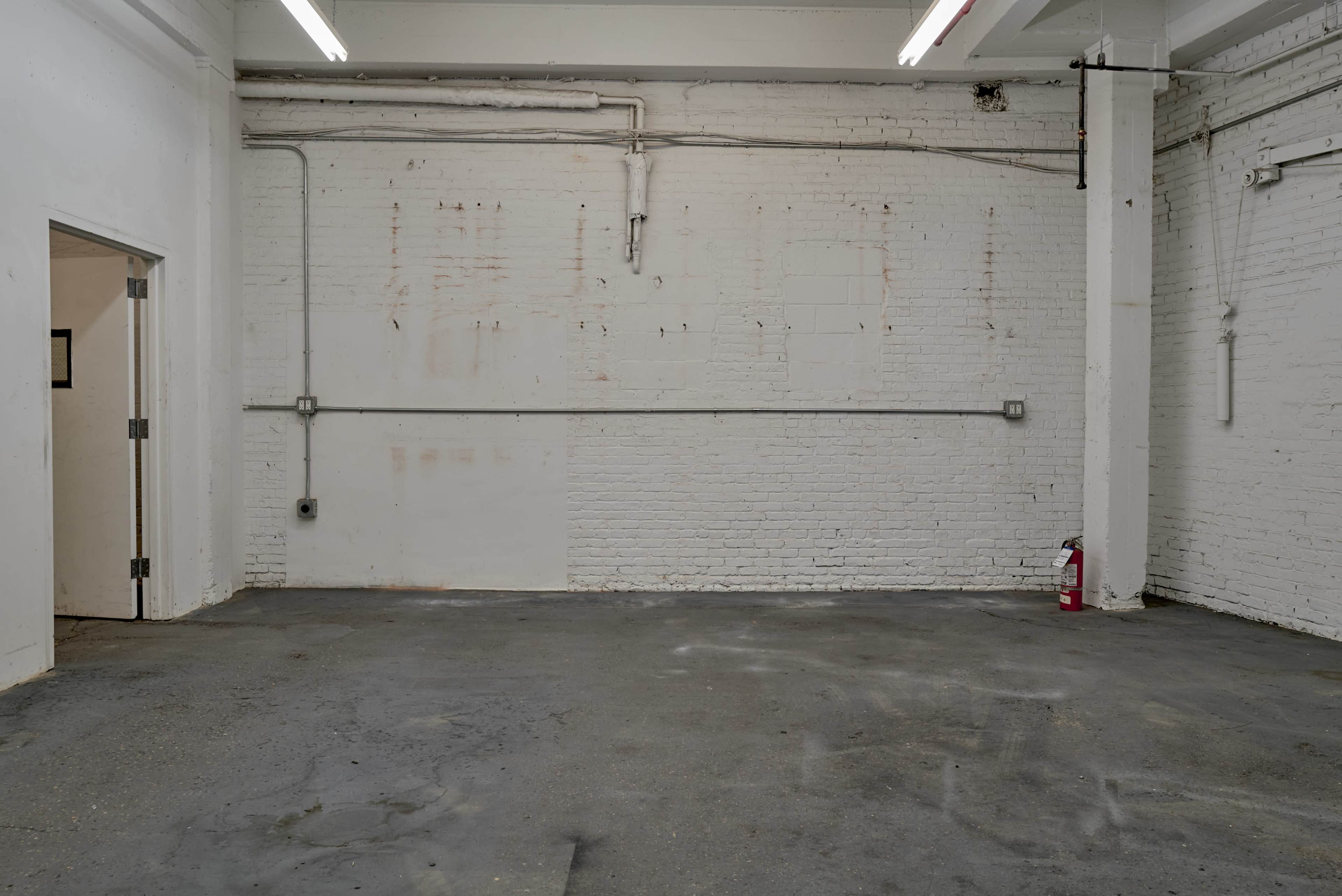 The image shows an empty, industrial-style room with bare white brick walls and a concrete floor.