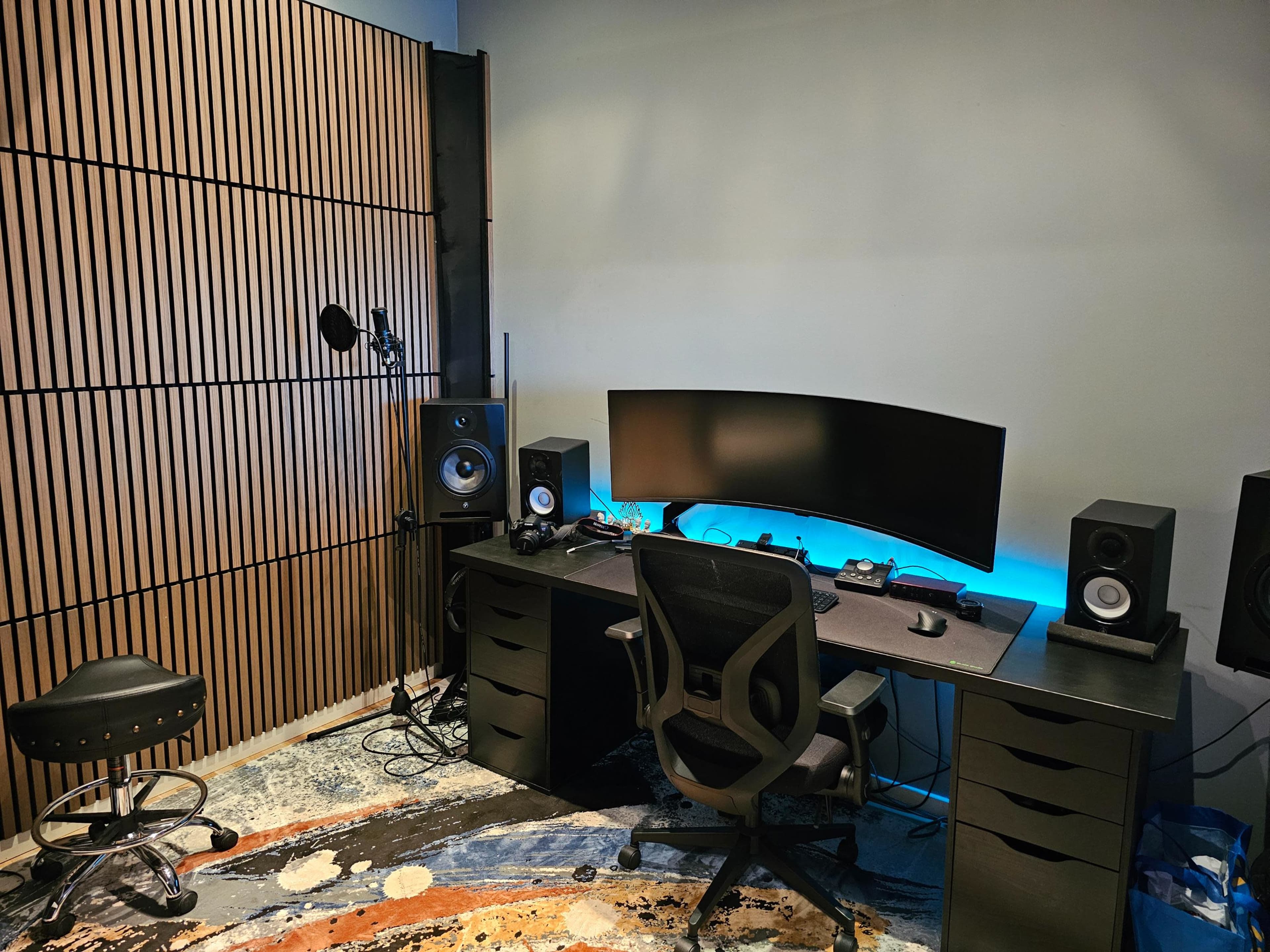 The image shows a modern workspace featuring a curved monitor, dual speakers, a black desk with storage drawers, an ergonomic chair, and a wooden accent wall.