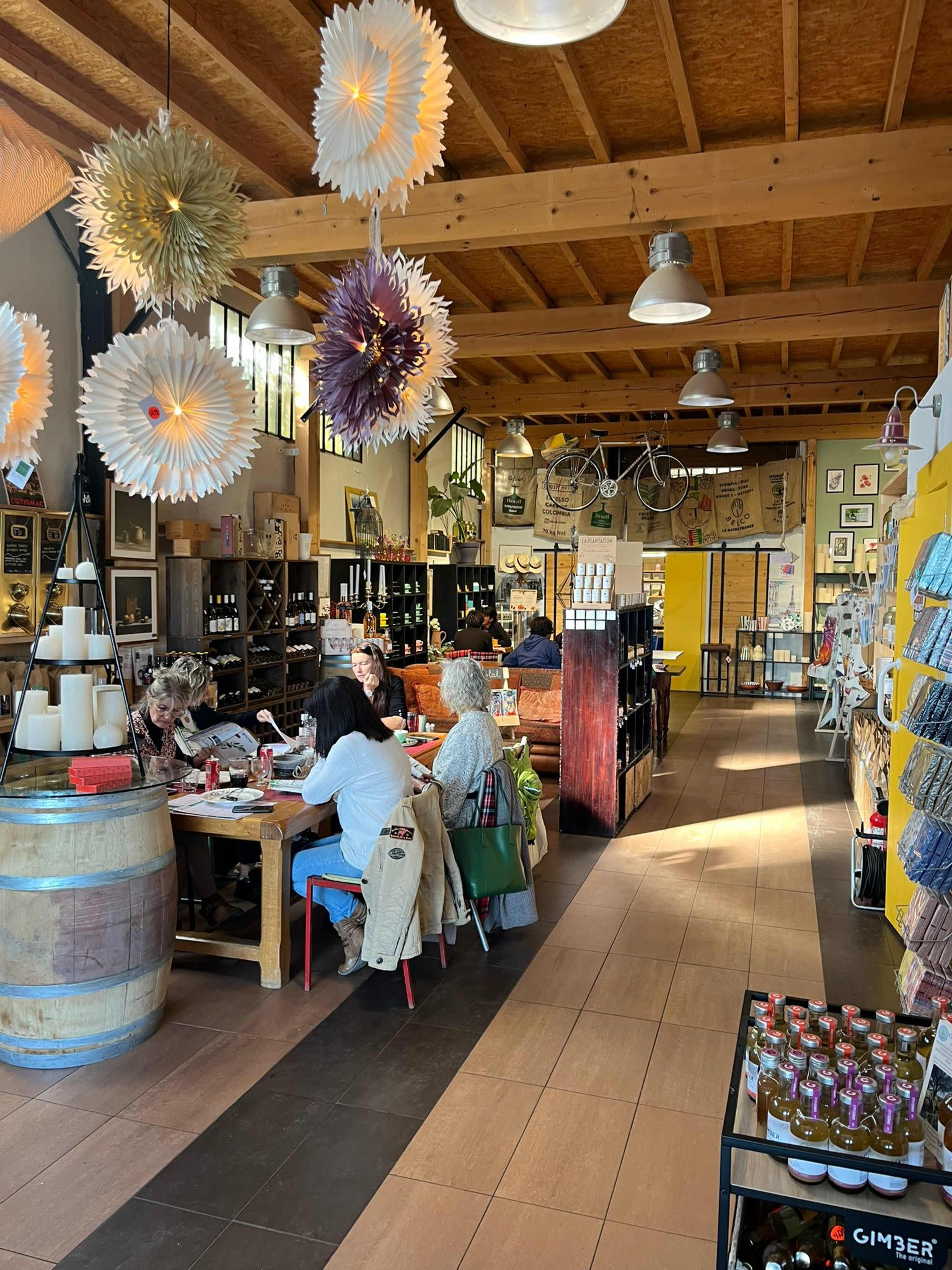 The interior of a boutique store features wooden beams, decorative paper flowers, shelves filled with various products, and customers seated around a table engaged in conversation.