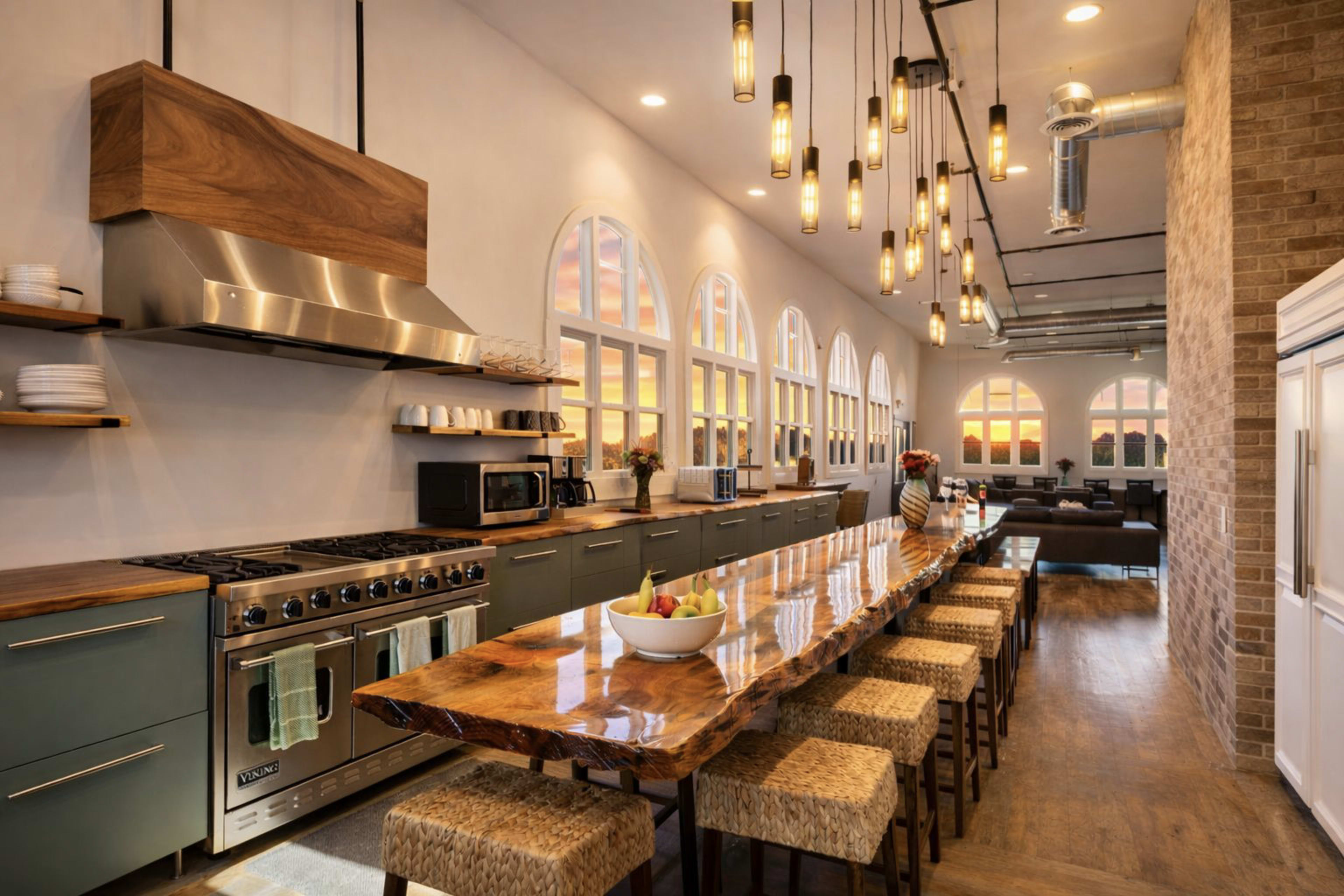 The image depicts a modern kitchen with a long wooden dining table, industrial light fixtures, and large windows showcasing a sunset.