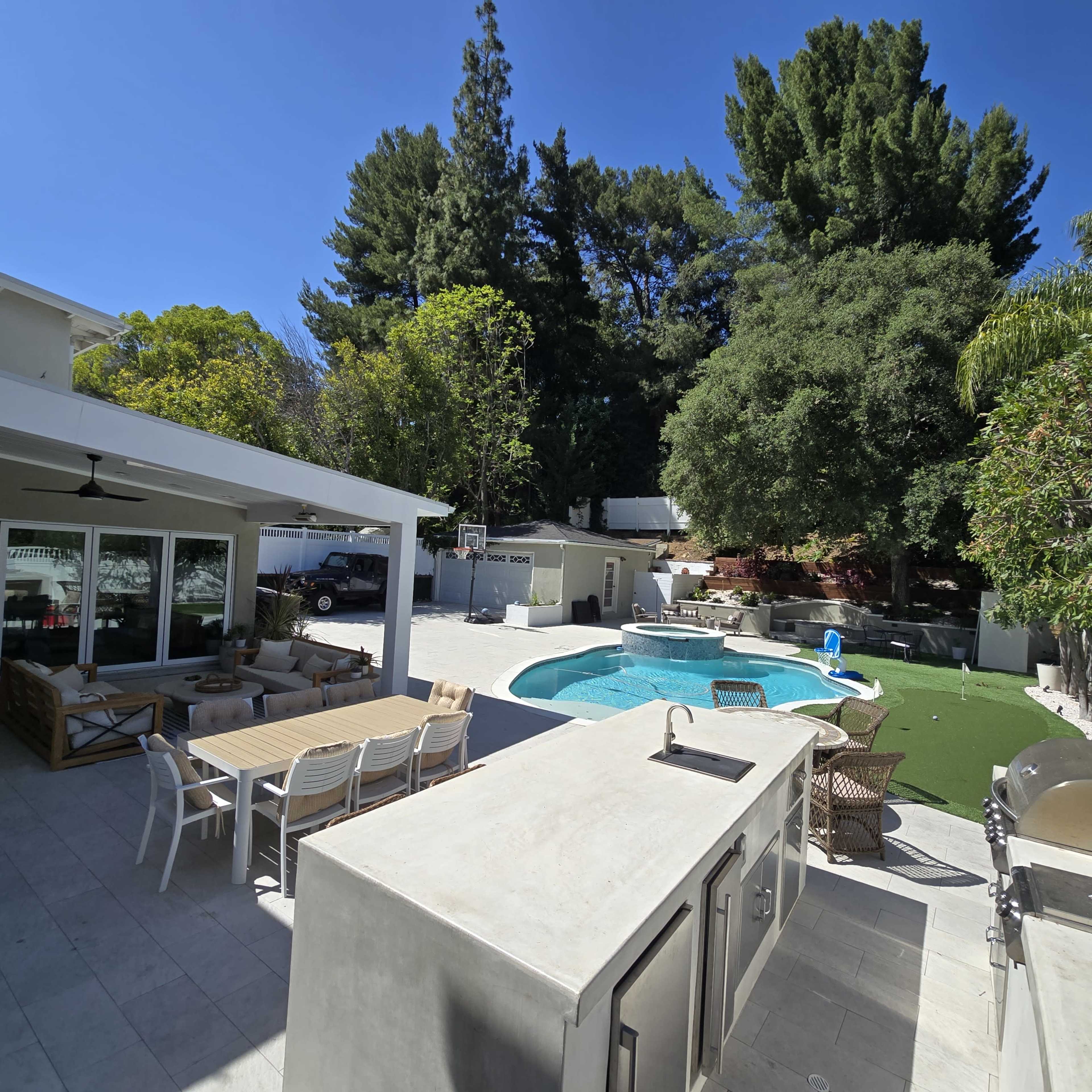 The image shows a spacious outdoor area with a pool, seating arrangements, and a barbecue grill surrounded by lush greenery and trees.