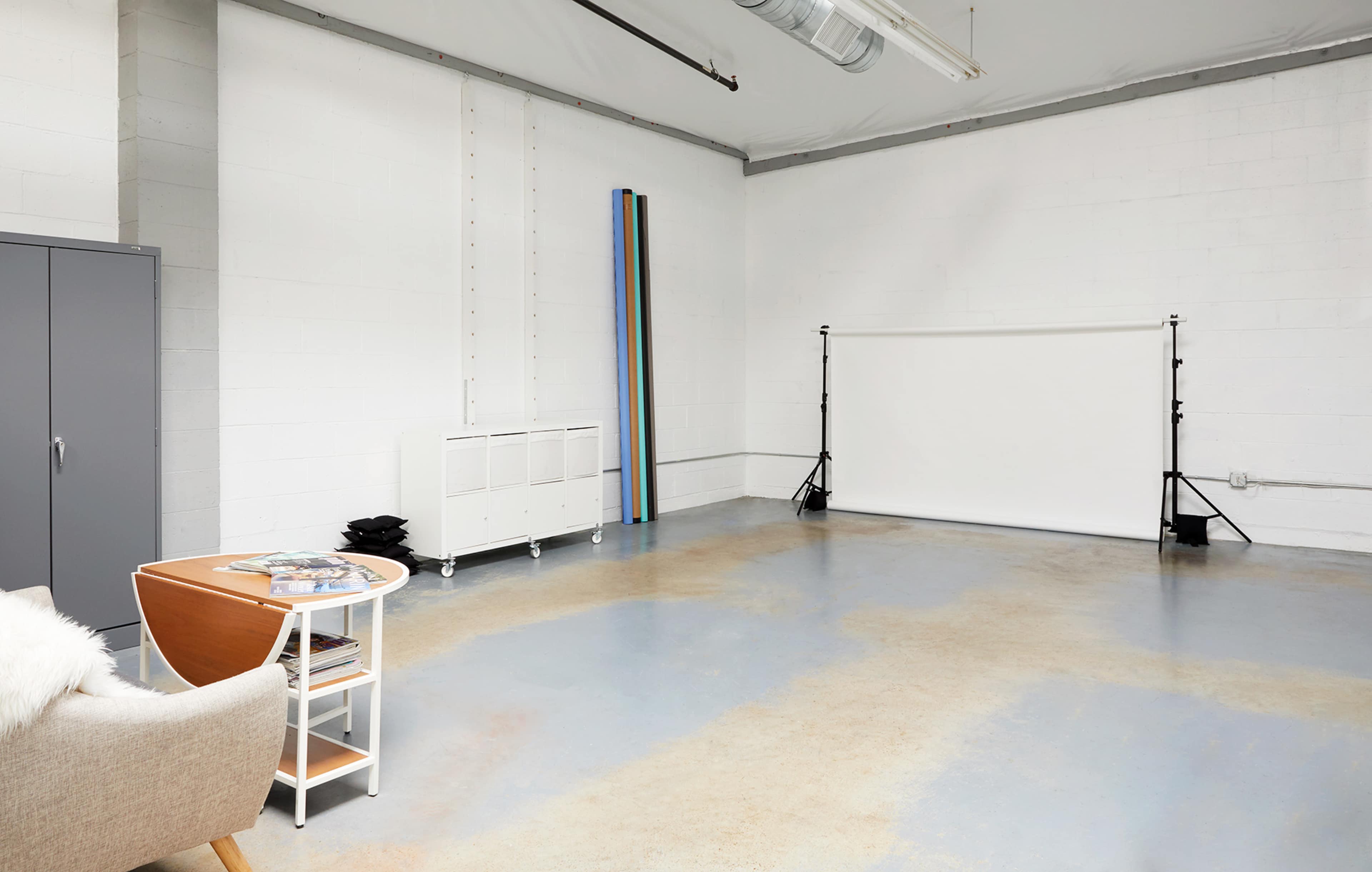 A spacious, empty studio with light gray walls and a white backdrop set up on stands, accompanied by a small table and a cabinet.