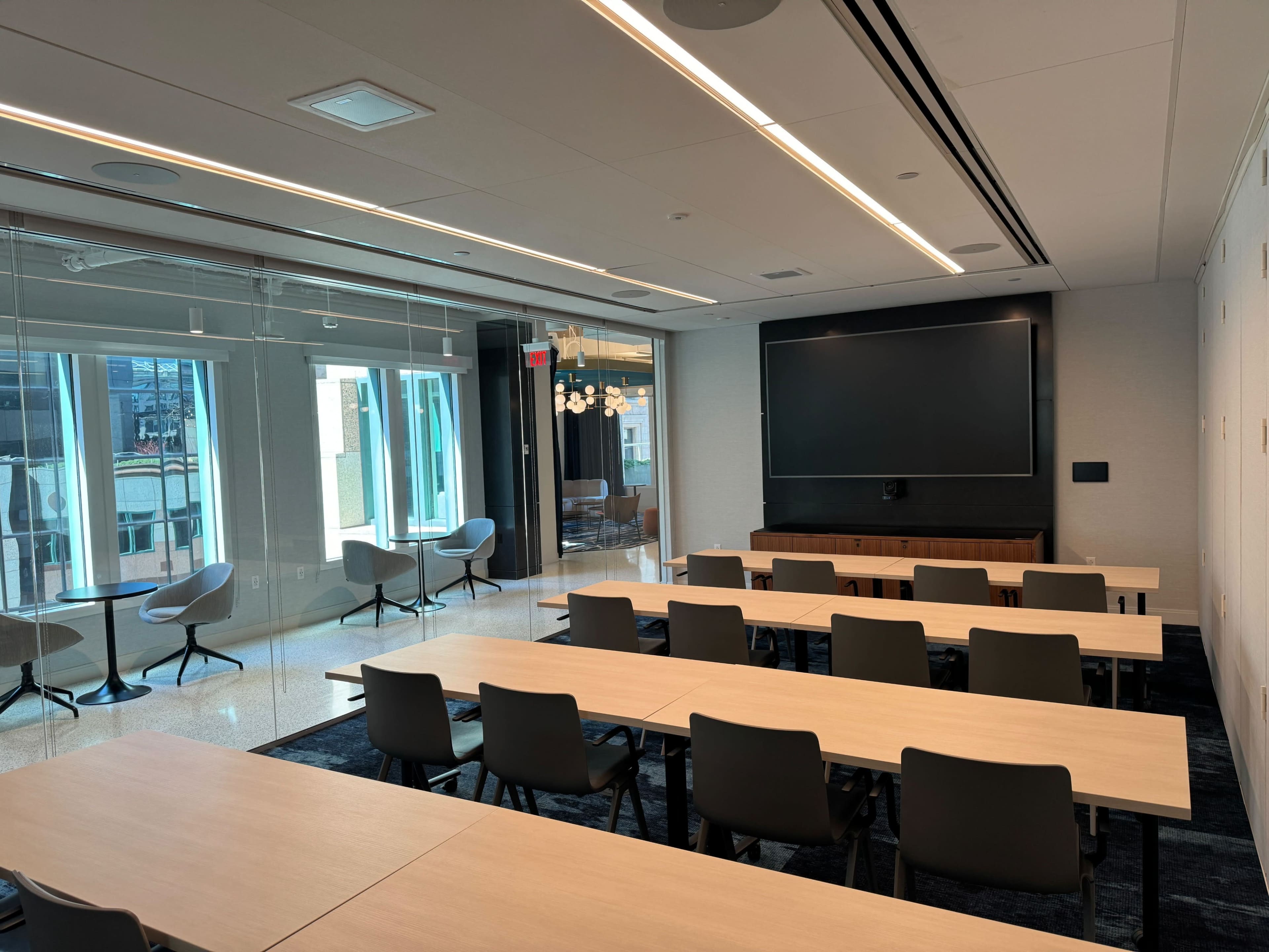 A modern conference room with rows of wooden tables and chairs facing a large screen at the front, surrounded by glass walls and windows.